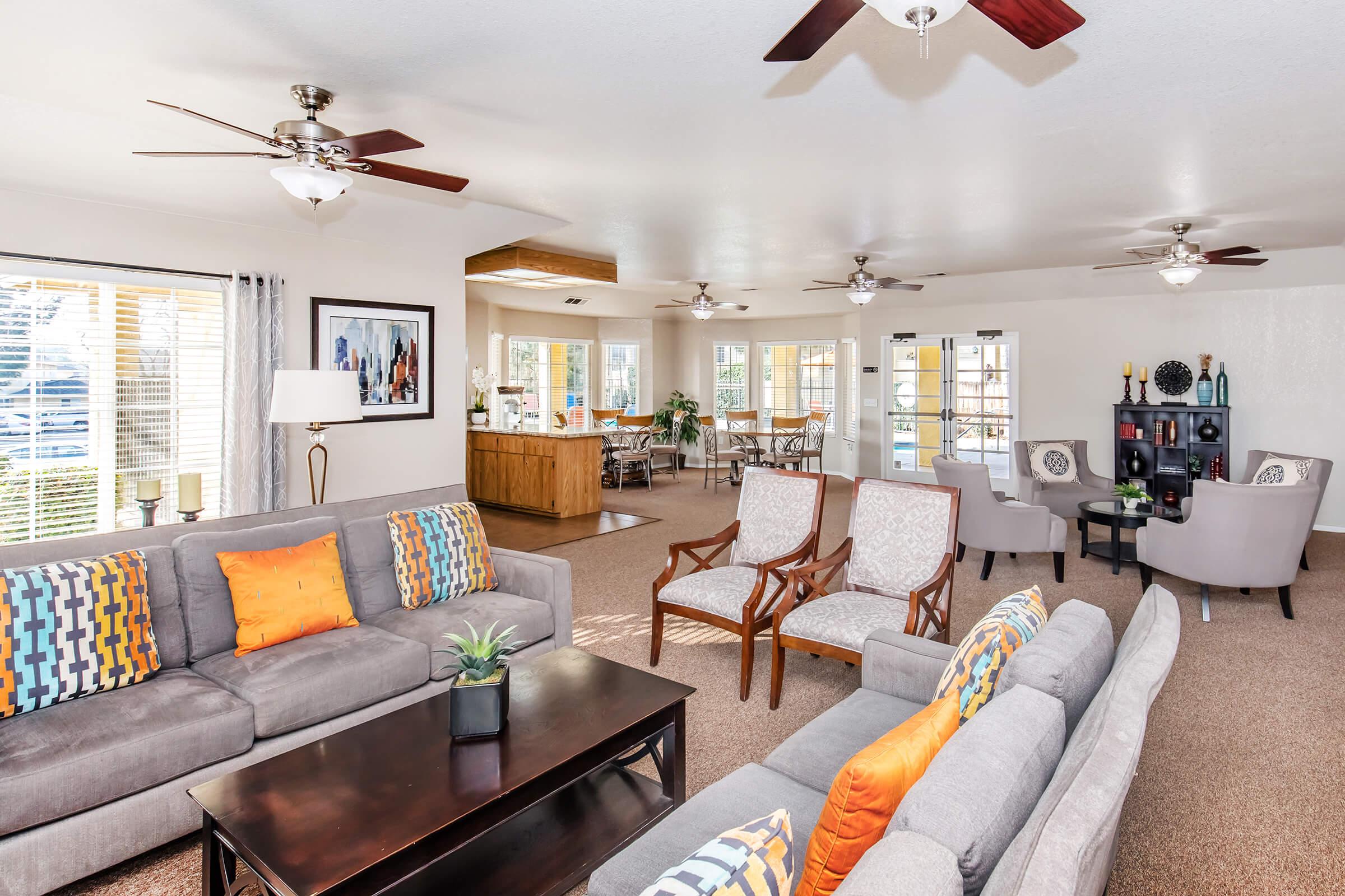 A spacious living room featuring two comfortable gray sofas with colorful cushions, a dark wooden coffee table, and stylish armchairs. The room is well-lit with ceiling fans and large windows, showcasing a warm and inviting atmosphere. In the background, a dining area with a wooden table can be seen.
