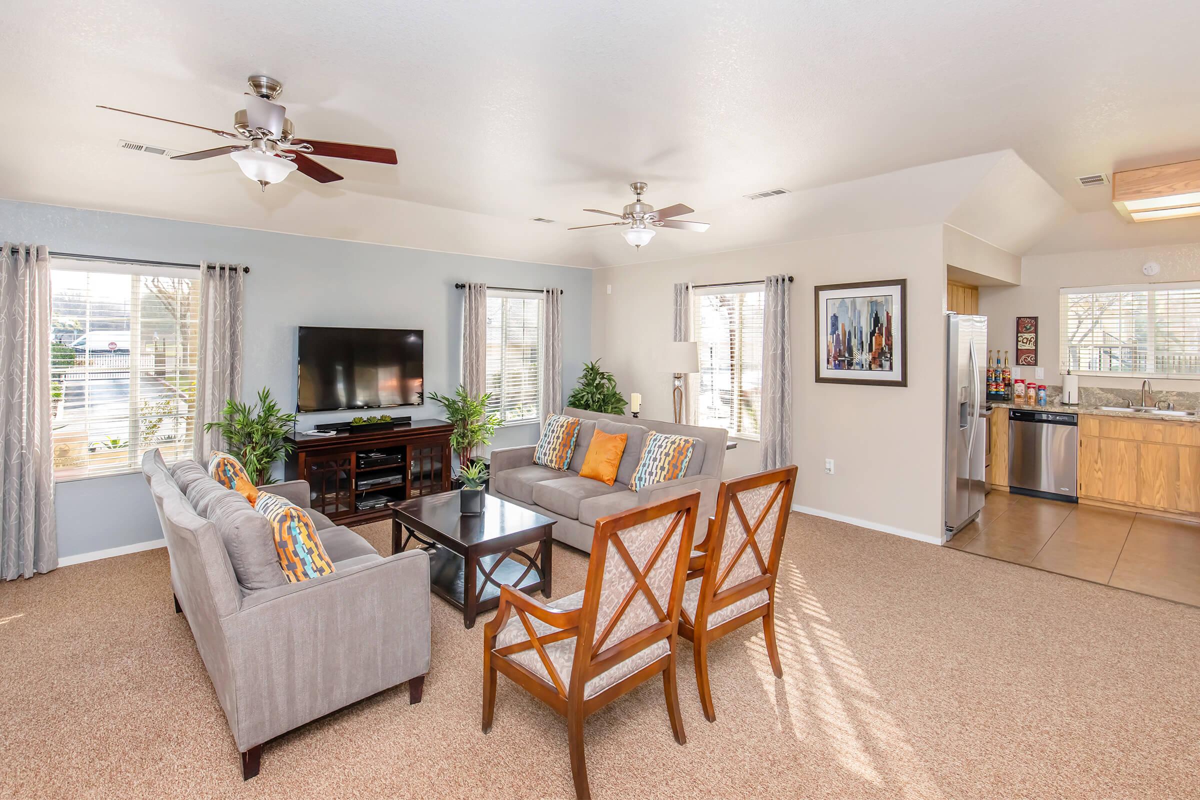 A bright and spacious living room featuring a comfortable gray sofa with colorful cushions, two wooden chairs, a flat-screen TV on a wooden stand, large windows with curtains, and a view of an open kitchen with modern appliances and tile flooring.