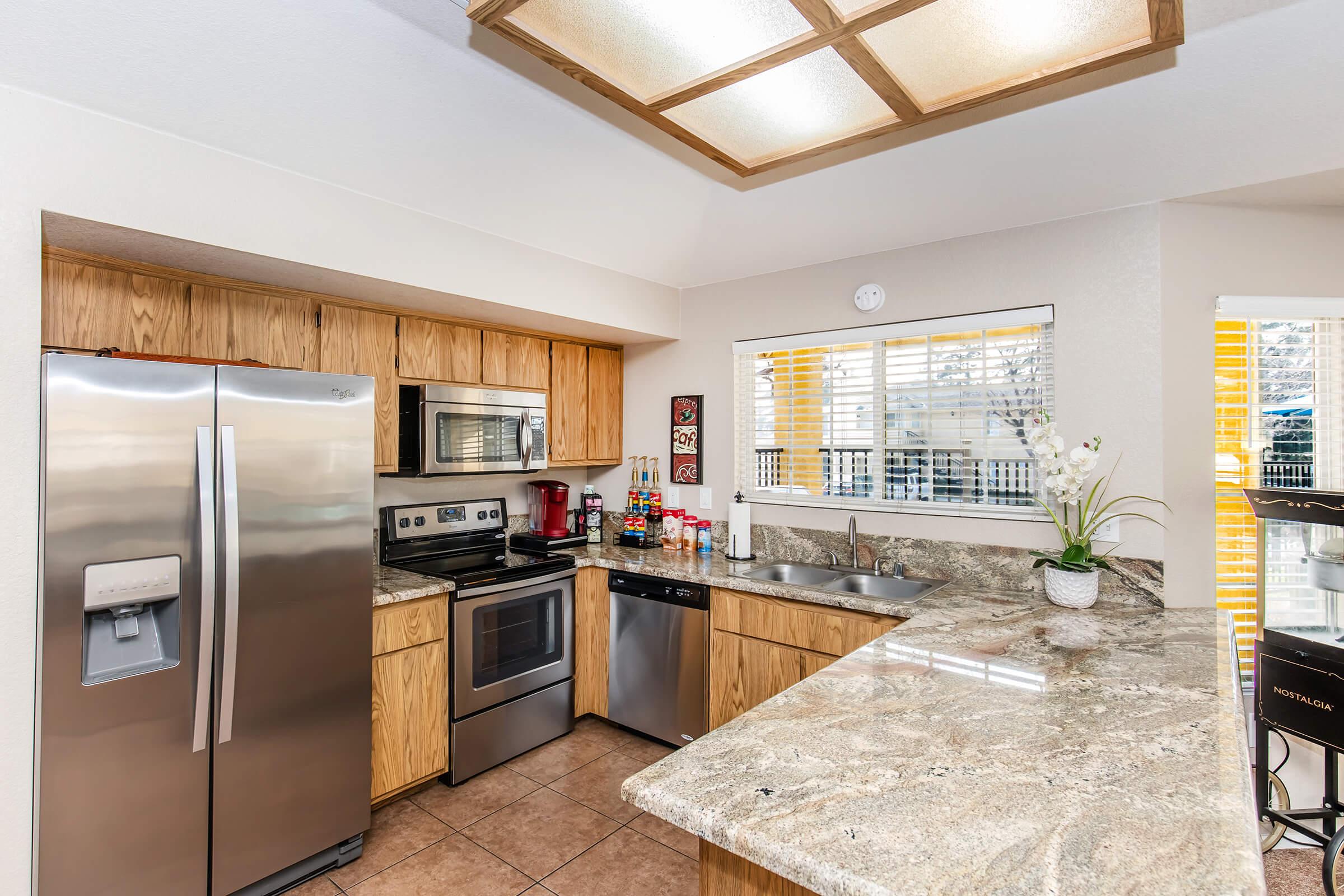 Modern kitchen featuring wooden cabinets, stainless steel appliances, granite countertops, and a tile floor. Includes a refrigerator, oven, microwave, dishwasher, and a sink with a window above. The space is well-lit with natural light and decorated with small plants and kitchenware.