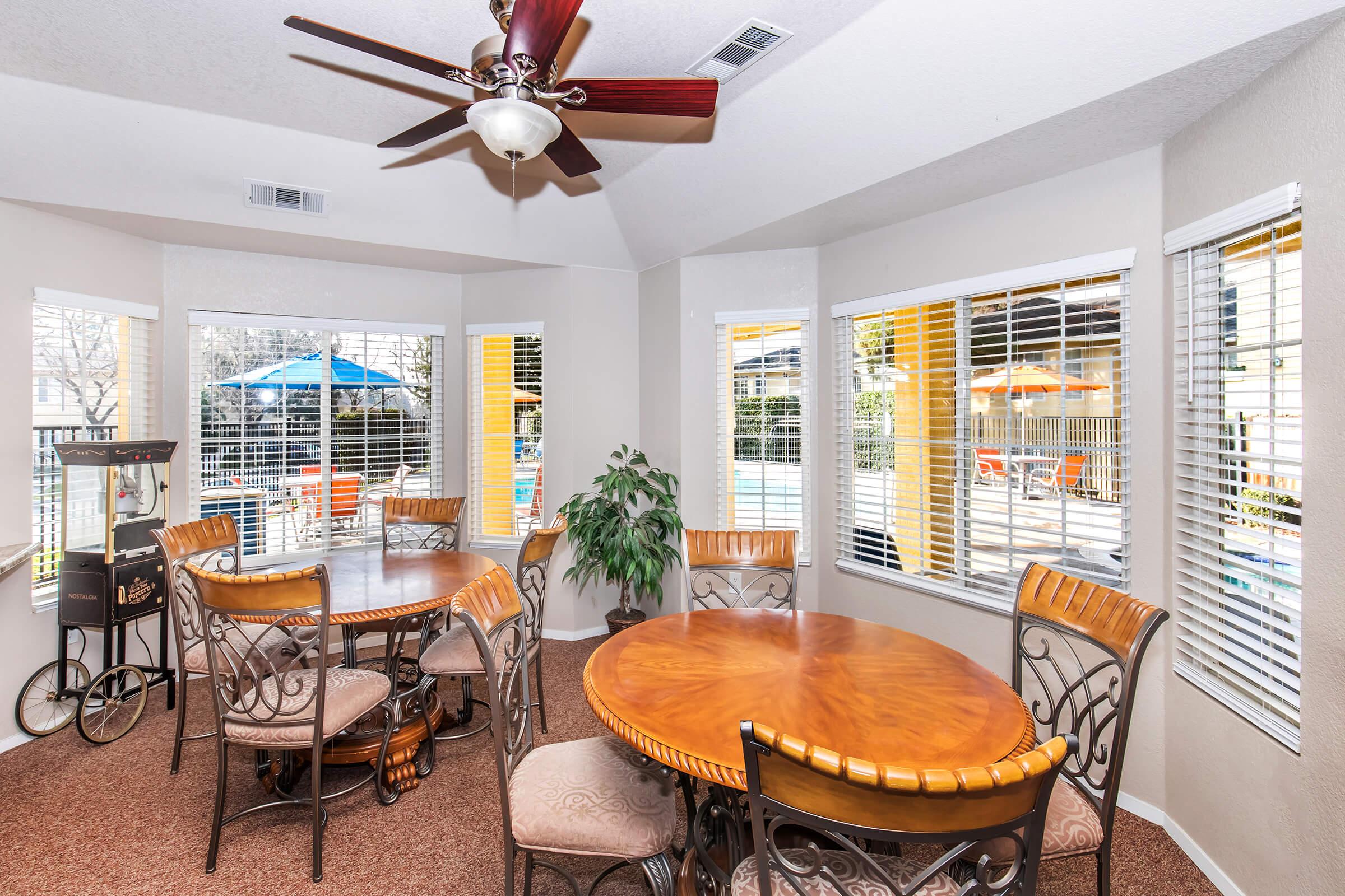 Bright dining area featuring a circular wooden table with six ornate chairs. Large windows allow natural light, showcasing a view of an outdoor pool area with blue umbrellas. A ceiling fan adds a cozy touch to the room, which is decorated with a potted plant and has a warm, inviting atmosphere.