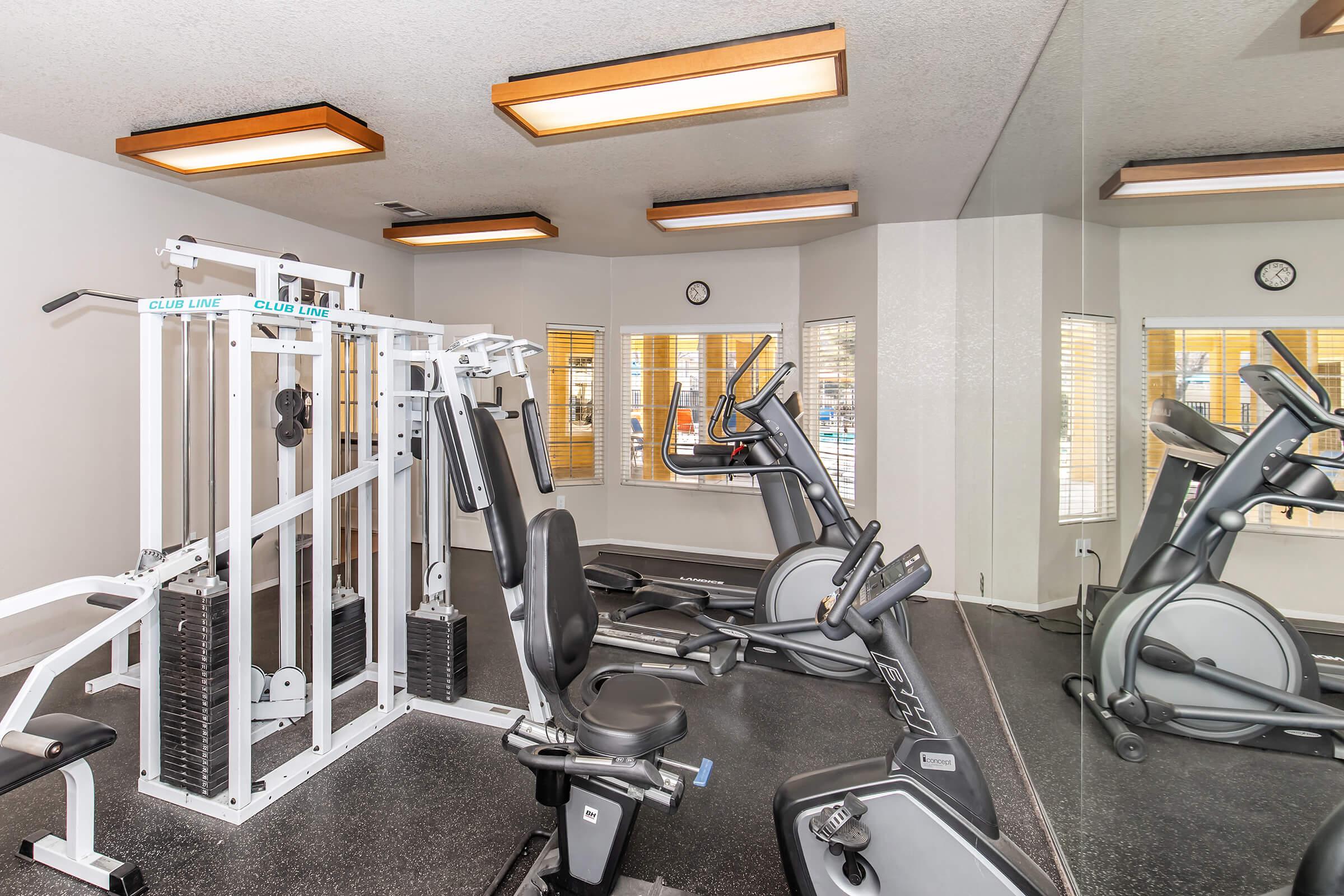 A gym interior featuring various exercise equipment, including a multi-gym machine, stationary bikes, and ellipticals. The space is well-lit with ceiling lights and has large windows allowing natural light. A mirror runs along one wall, enhancing the sense of openness in the room.