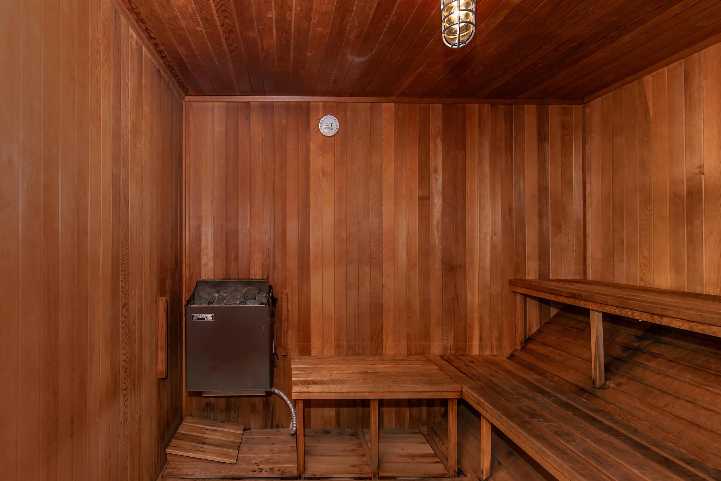 A wooden sauna interior featuring smooth wooden walls and benches. There is a metal stove in the corner, a clock on the wall, and soft lighting from a hanging fixture. The space is designed for relaxation and heat therapy, showcasing a warm and inviting atmosphere.