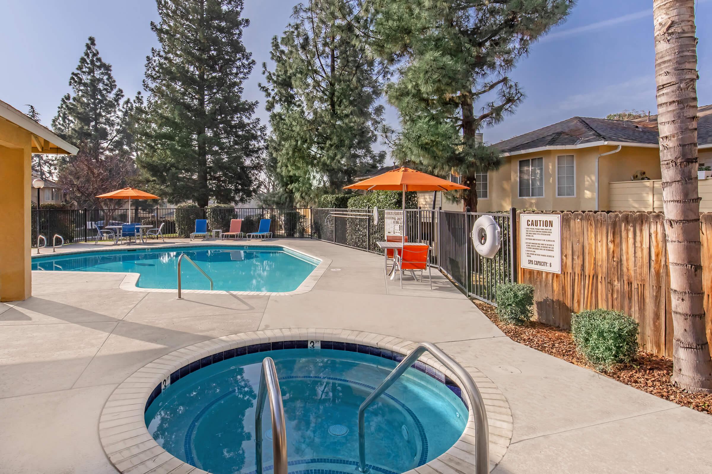 Swimming pool area featuring a large pool and a hot tub. The pool is surrounded by lounge chairs and orange umbrellas. Tall trees provide shade, and there's a wooden fence in the background. A sign indicating caution is visible near the hot tub. Bright, sunny day with clear blue skies.