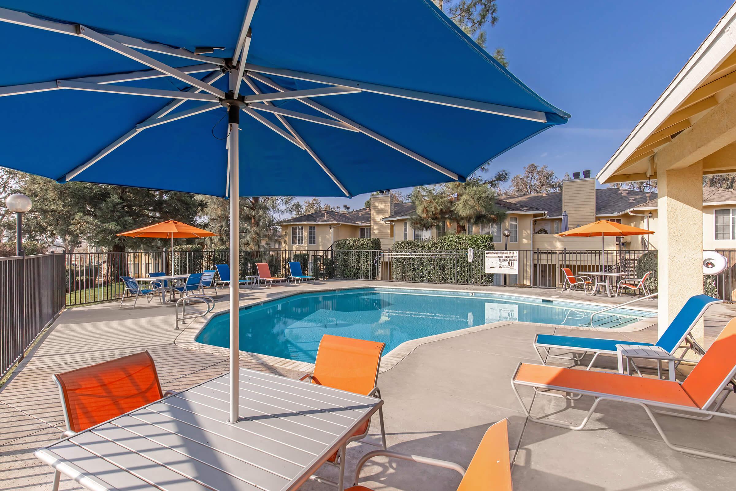 A bright and inviting outdoor swimming pool area featuring a clear blue pool surrounded by lounge chairs in orange and blue, umbrellas providing shade, and a well-maintained landscape. The area is fenced for privacy, enhancing the relaxing atmosphere ideal for leisure and recreation.