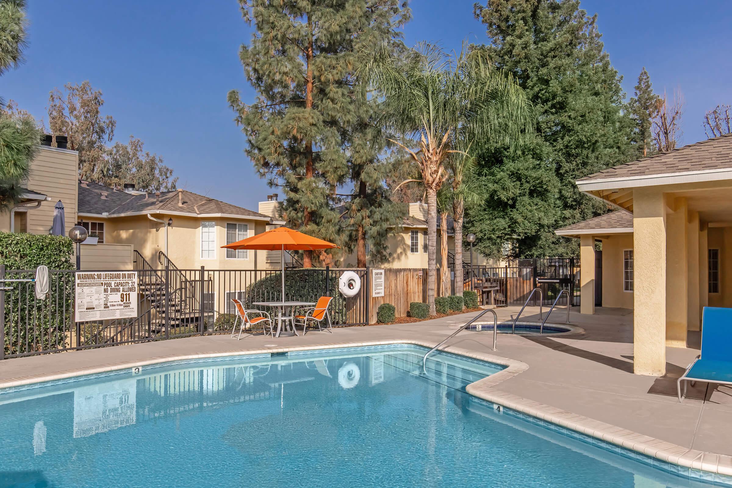 A swimming pool surrounded by lounge chairs and an orange umbrella, with greenery and trees in the background. Adjacent buildings are visible, along with a safety sign near the pool area, creating a relaxing outdoor environment.