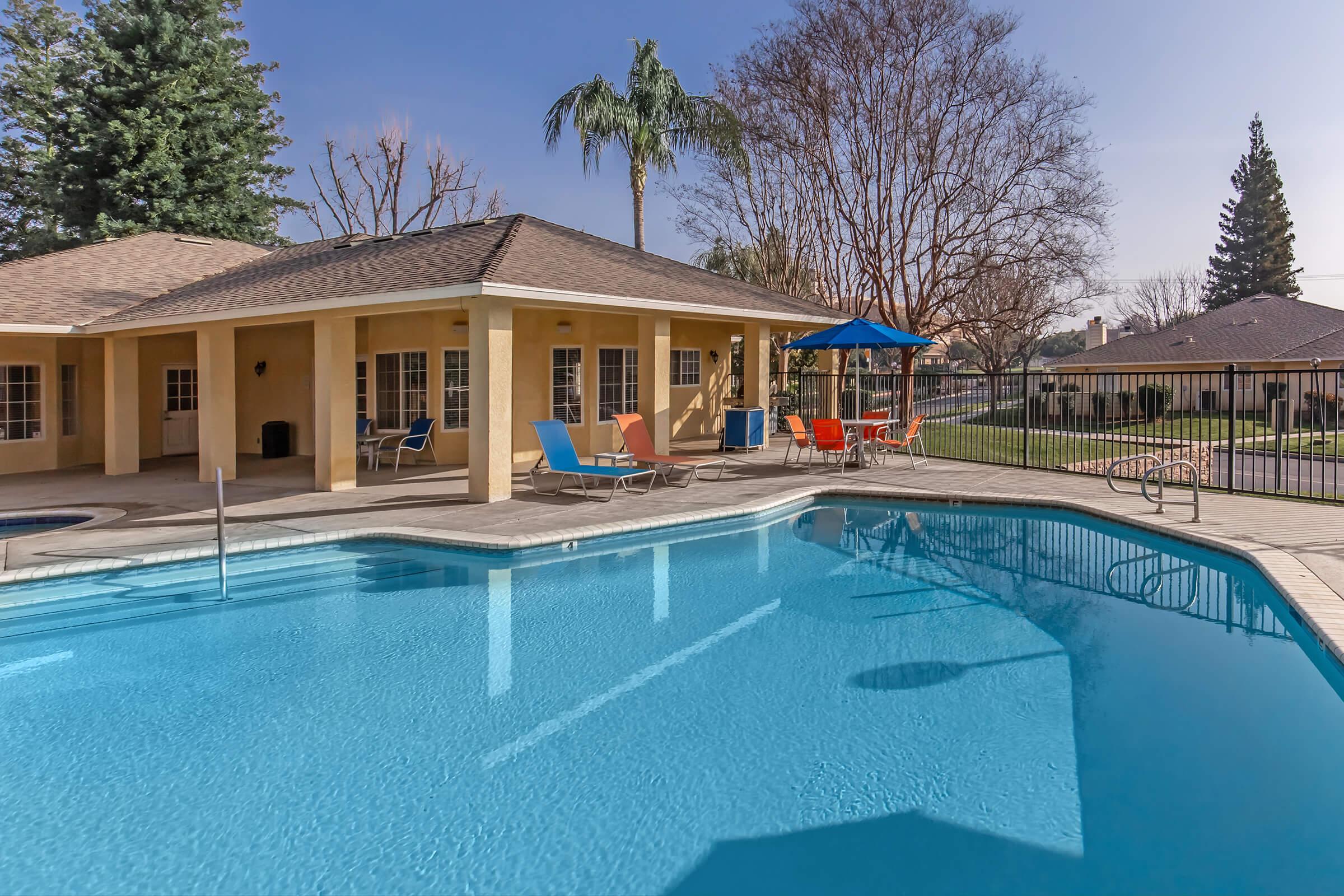 A clear blue swimming pool surrounded by a patio with colorful lounge chairs. In the background, there are trees and a fenced area, along with a single-story house featuring a tan exterior. Bright sunlight enhances the inviting atmosphere of the outdoor space.