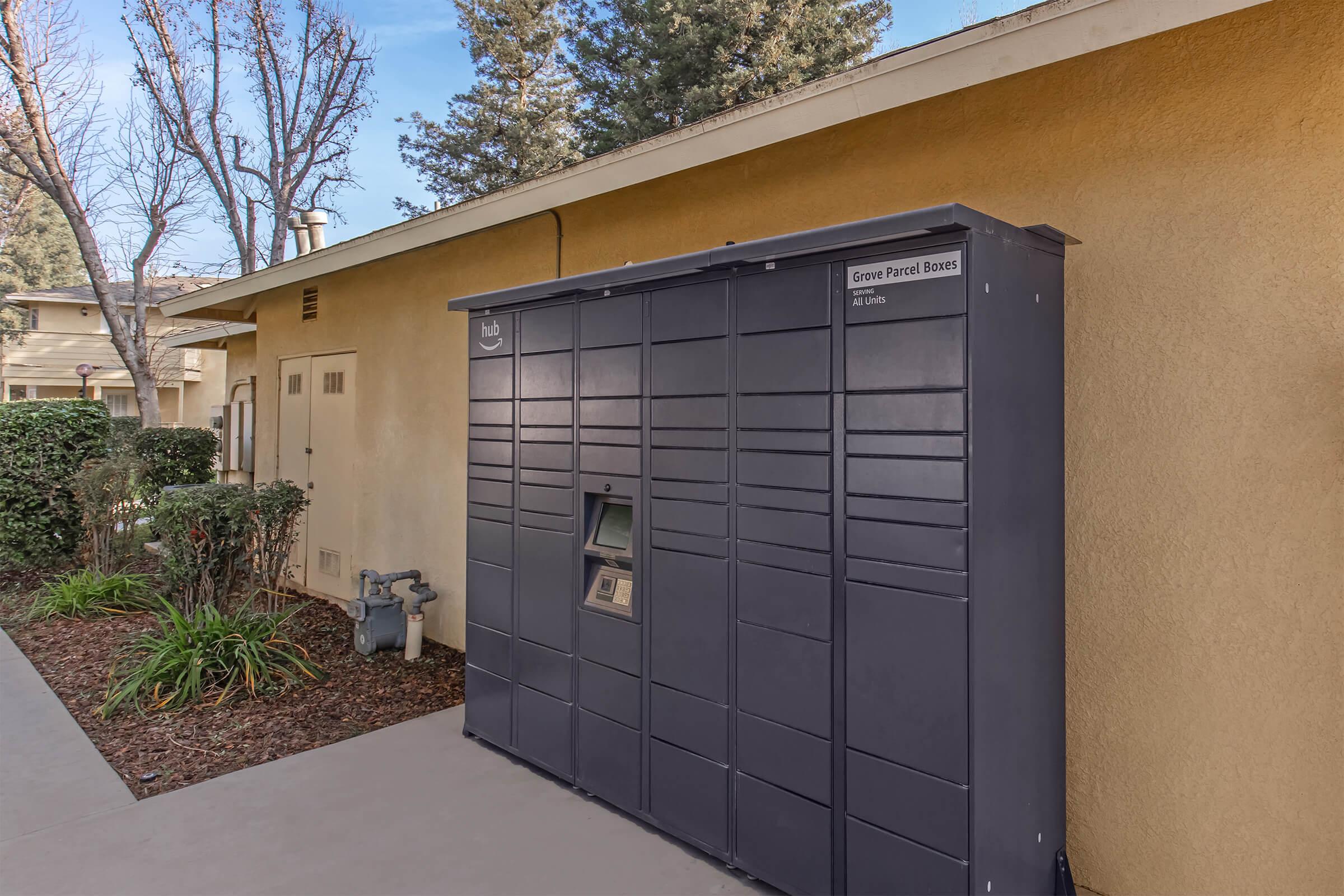 A large, dark-colored parcel locker stationed next to a building, surrounded by low shrubs and trees. The locker features multiple compartments for package delivery and a small screen or interface for user interaction. The setting appears to be a residential area with soft daylight.