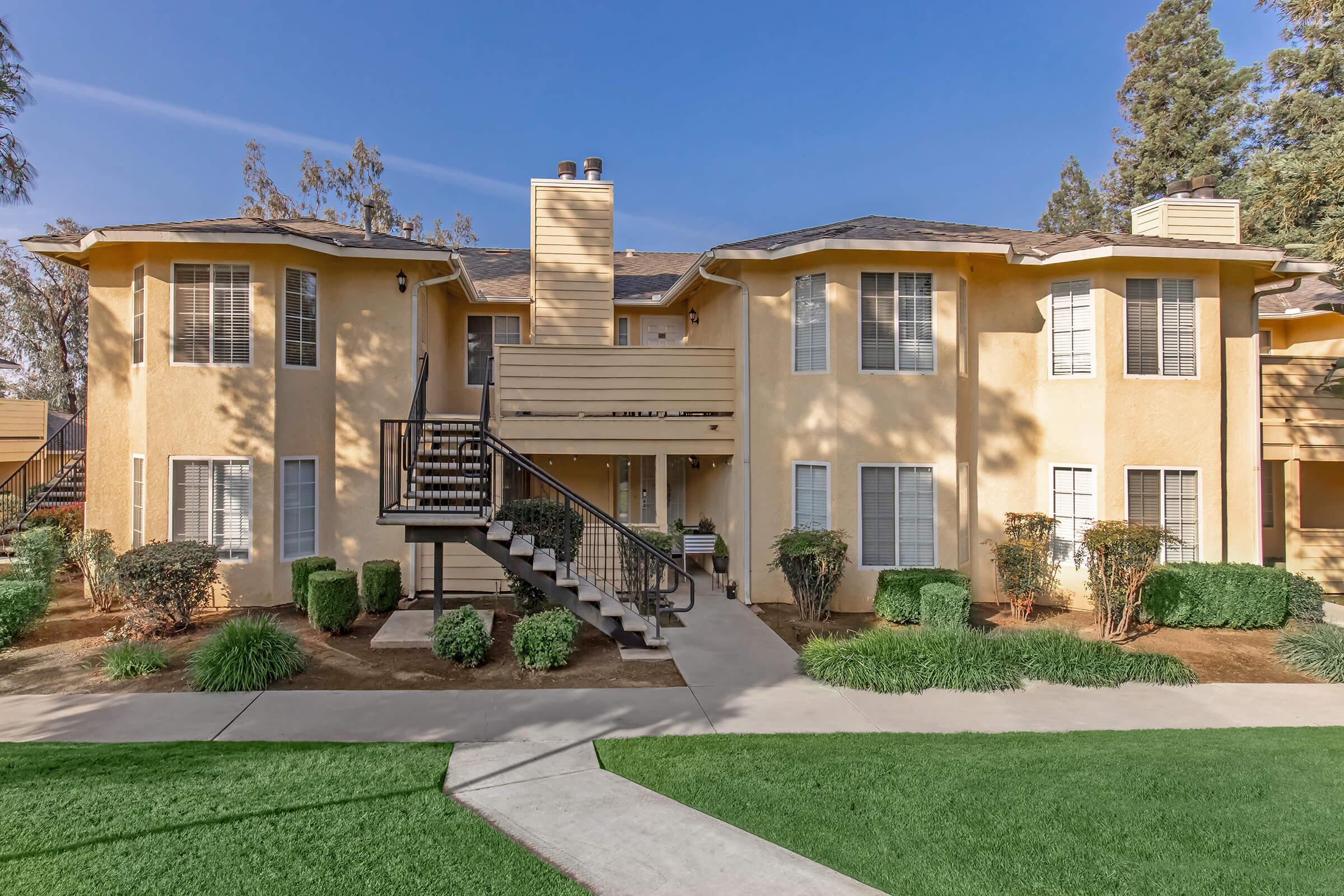 Two-story apartment building with beige exterior, surrounded by green lawns and shrubs. A set of stairs leads to the entrance on the left side, with several windows visible. The sky is clear and blue, enhancing the bright appearance of the structural design.