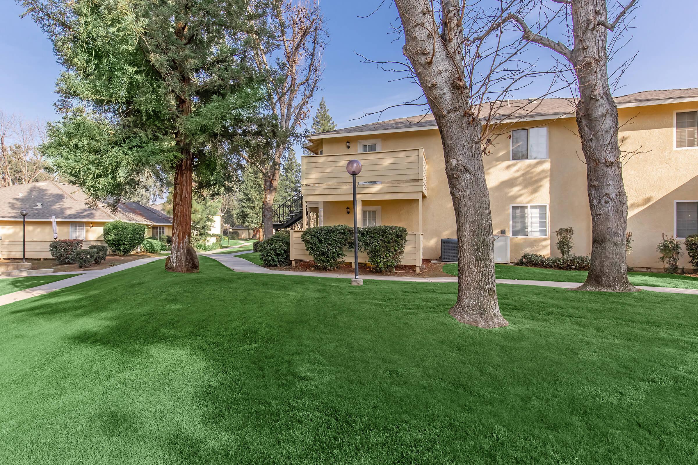 A landscaped area featuring well-maintained green grass, two trees, and light posts, with residential buildings in the background. The scene is bright and sunny, showcasing a peaceful apartment complex setting with clear blue skies.