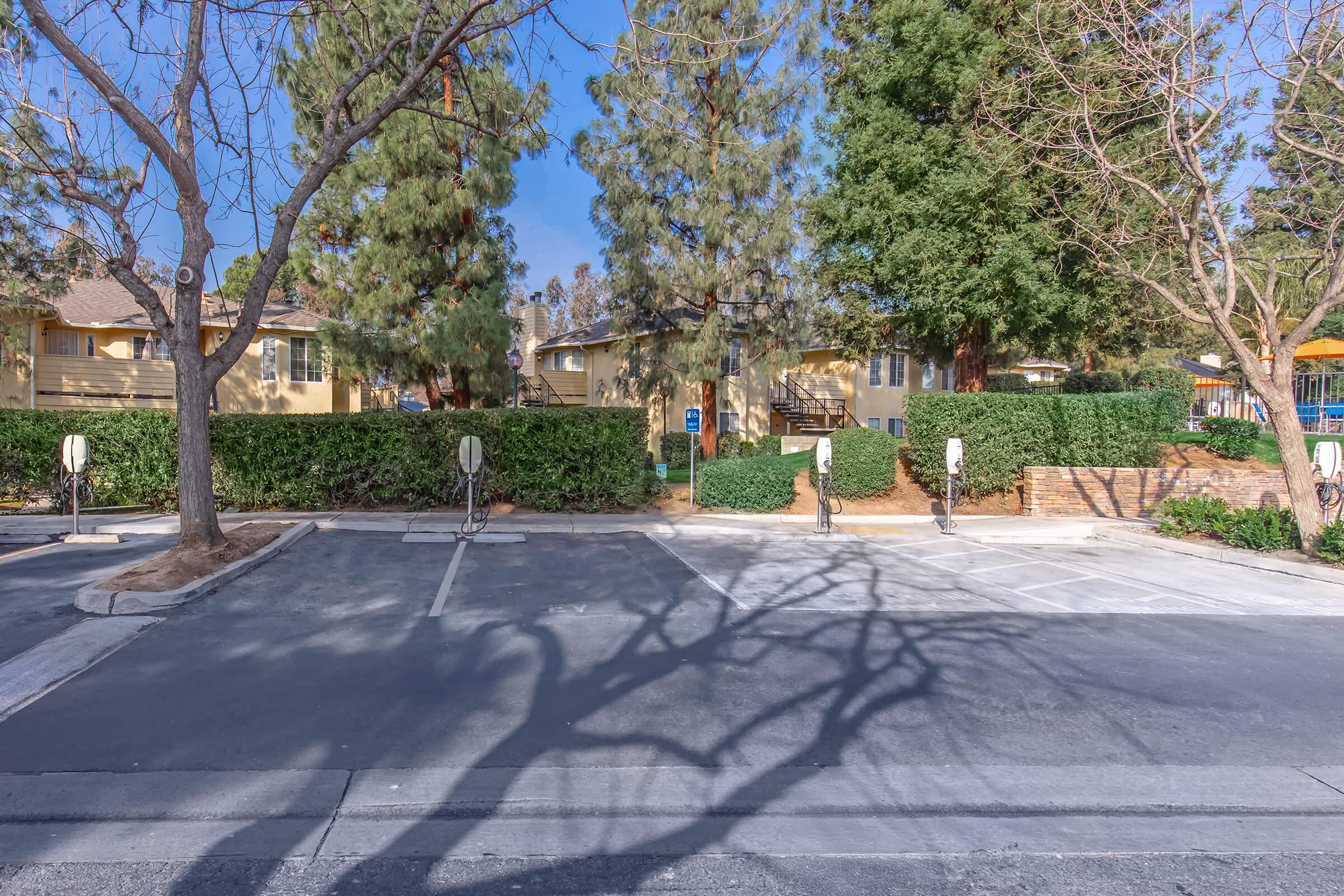 A parking lot with several empty spaces. Shadow patterns from surrounding trees are visible on the pavement. In the background, there are buildings partially obscured by greenery. The scene is clear and well-lit, with a blue sky overhead.