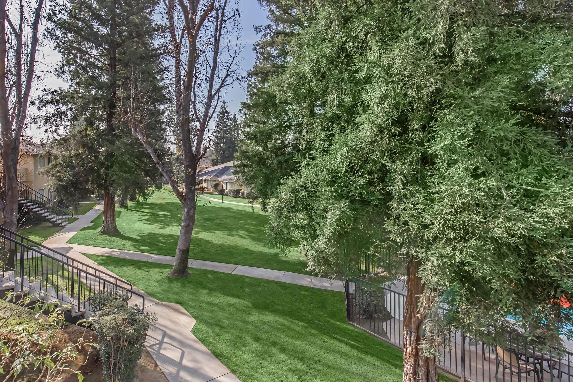A well-maintained green space featuring lush grass and tall trees. There are walkways winding through the area, leading to nearby structures. The setting appears peaceful, with clear skies and a residential atmosphere.