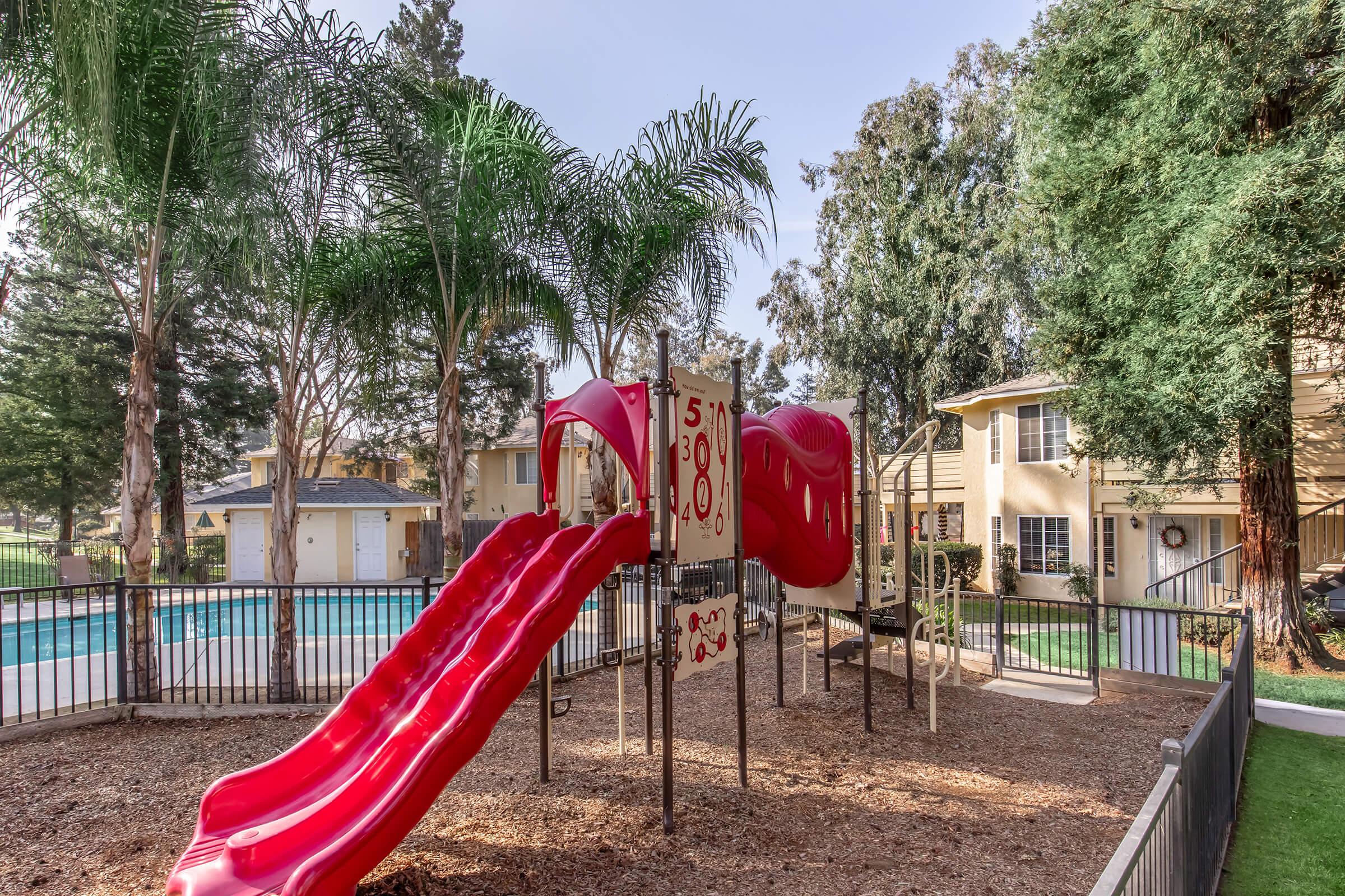 A playground with a red slide and climbing structure, surrounded by trees. In the background, there are residential buildings and a pool area. The ground is covered with wood chips. The setting appears sunny and inviting, perfect for children's outdoor play.