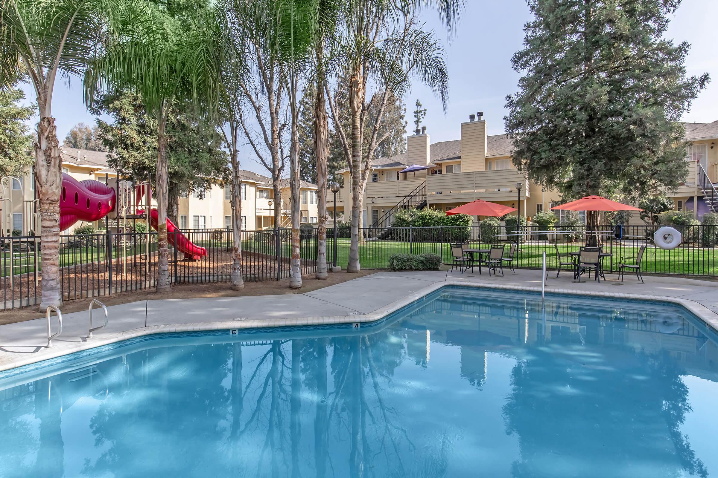 A sunny view of a swimming pool surrounded by tall palm trees and well-maintained landscaping. In the background, there is a playground with a slide and a grassy area with patio furniture and umbrellas, offering a relaxing outdoor environment for residents.