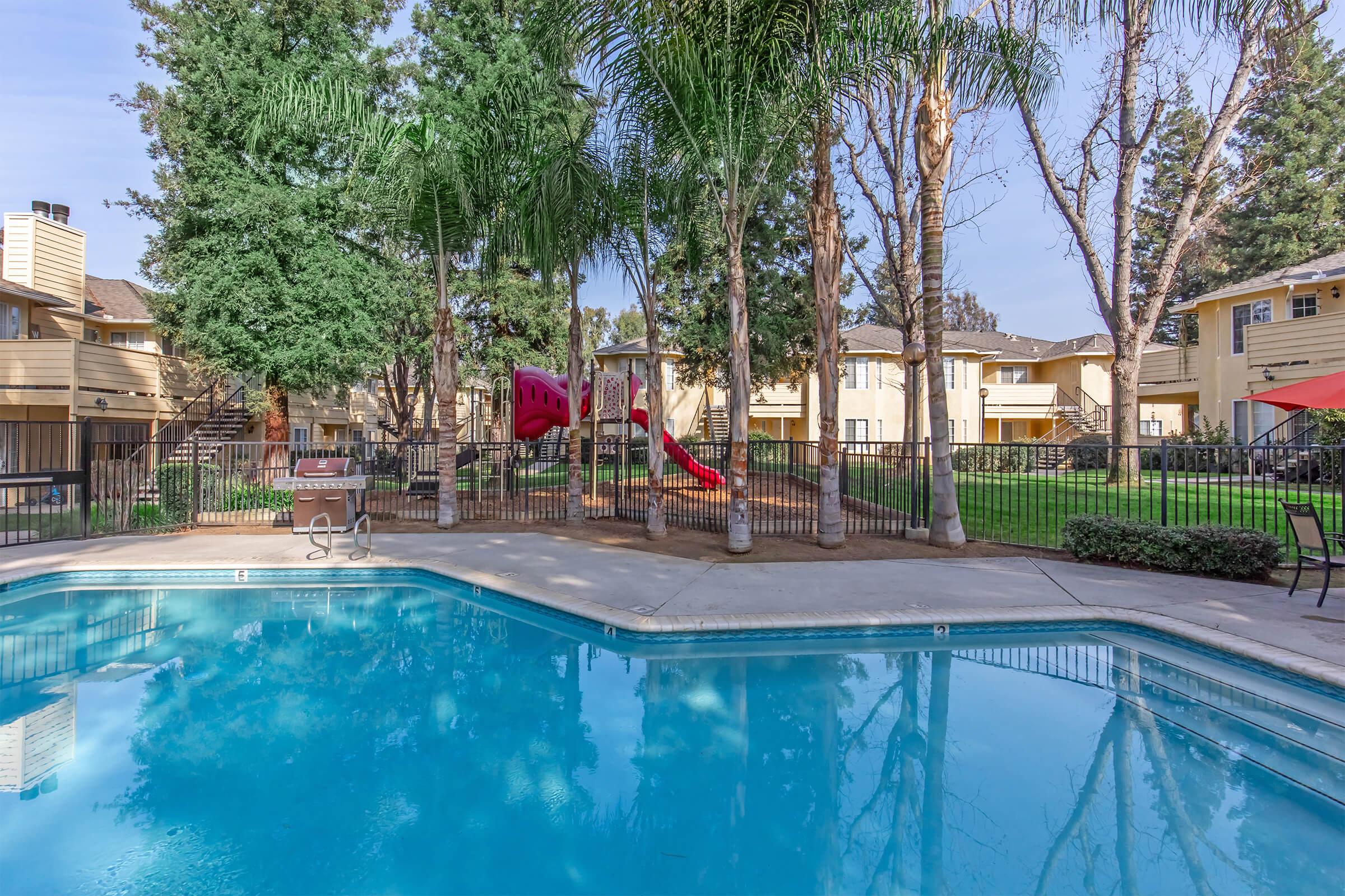 A residential community featuring a swimming pool with clear blue water, surrounded by palm trees and a grassy play area. In the background, there is a playground with a red slide, and several apartment buildings visible. The setting is bright and inviting, perfect for family activities.