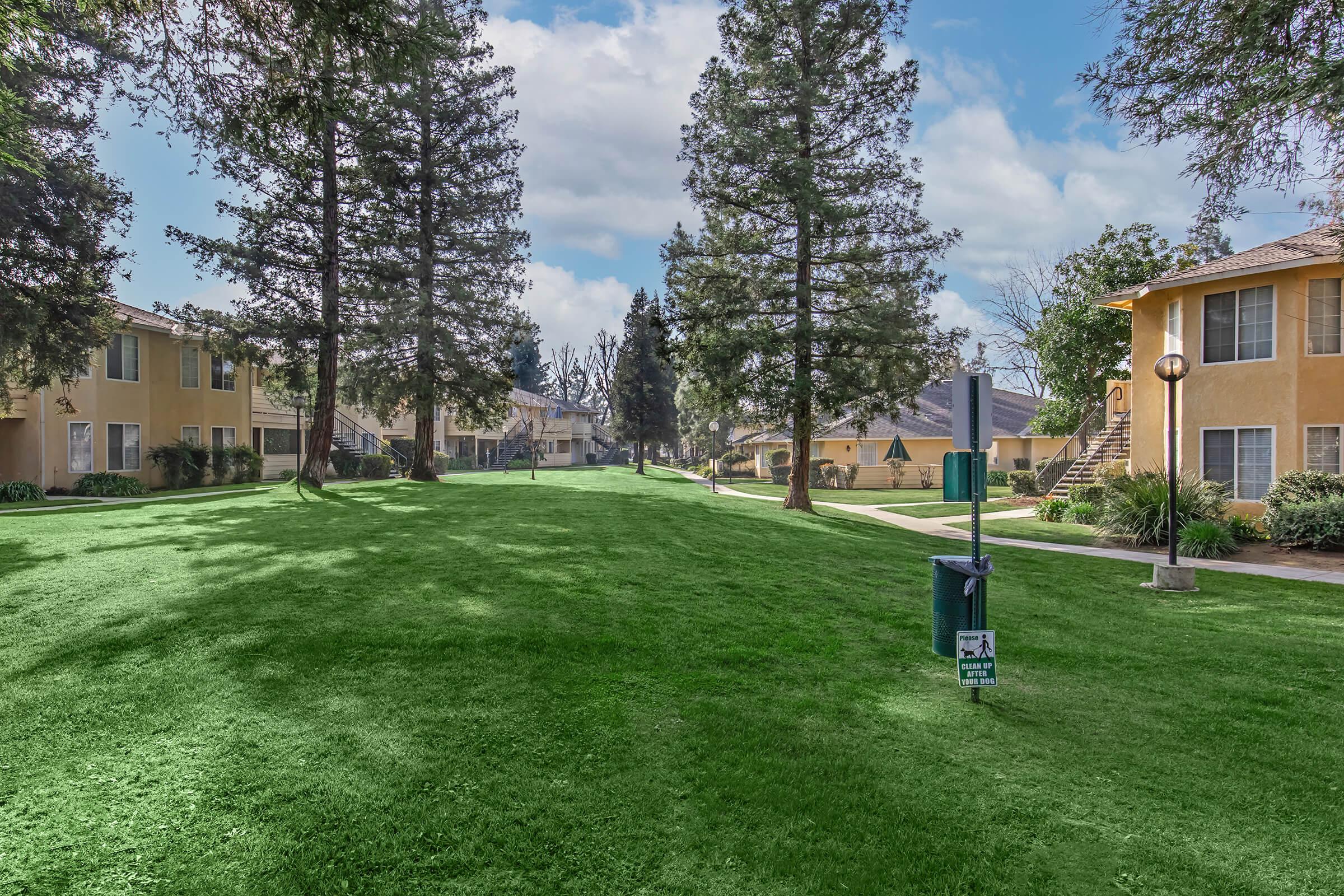 A green, well-maintained lawn bordered by two-story residential buildings with large windows. Tall trees provide shade, creating a peaceful outdoor space. A walking path runs through the area, with a trash bin and lighting fixtures visible. The scene is bright, indicating a clear day.