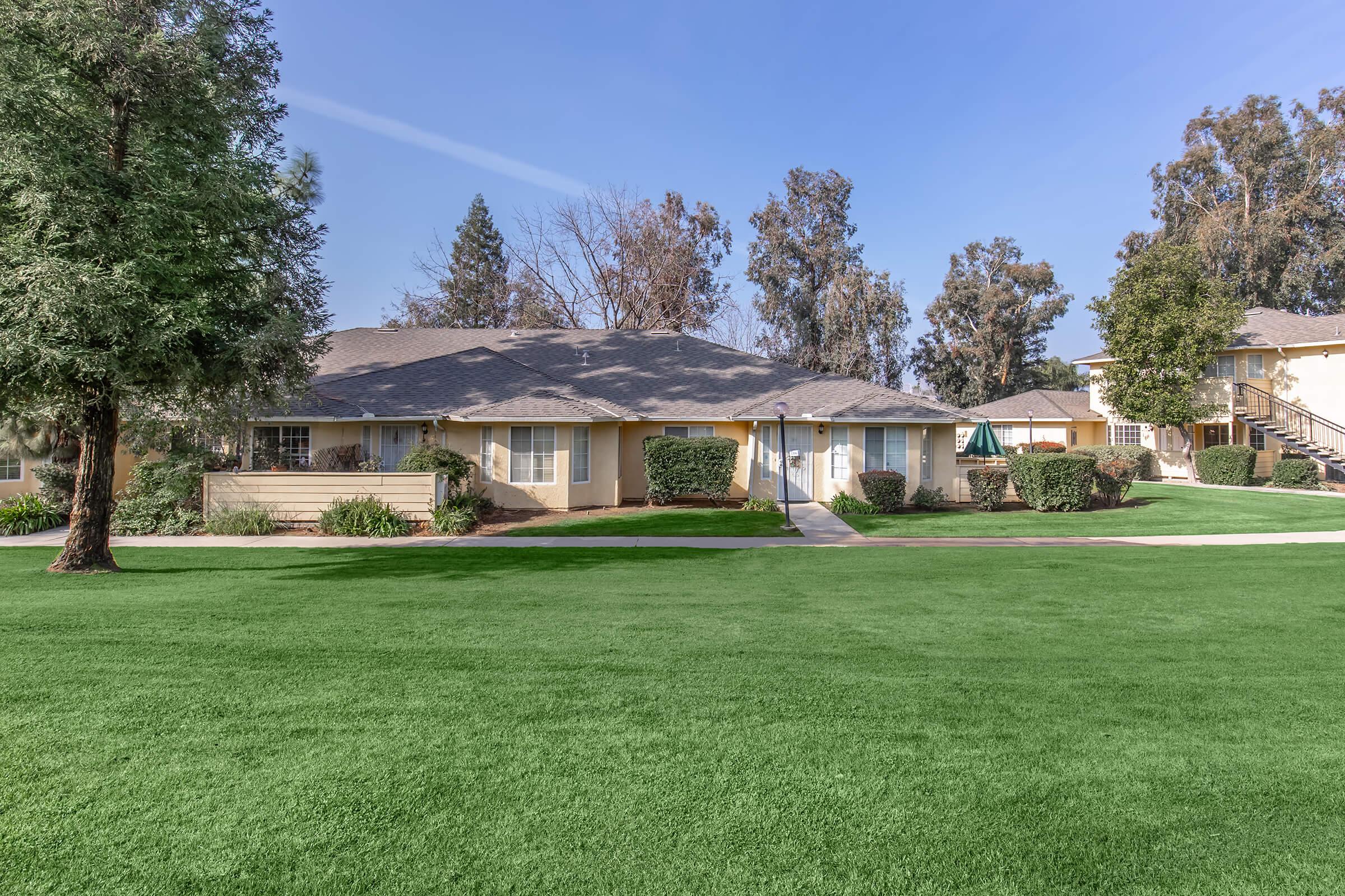 A well-maintained residential complex featuring single-story apartments surrounded by lush green lawns, trees, and shrubs. Clear blue sky overhead and a pathway leading to the entryways of the units. The setting appears peaceful and inviting.