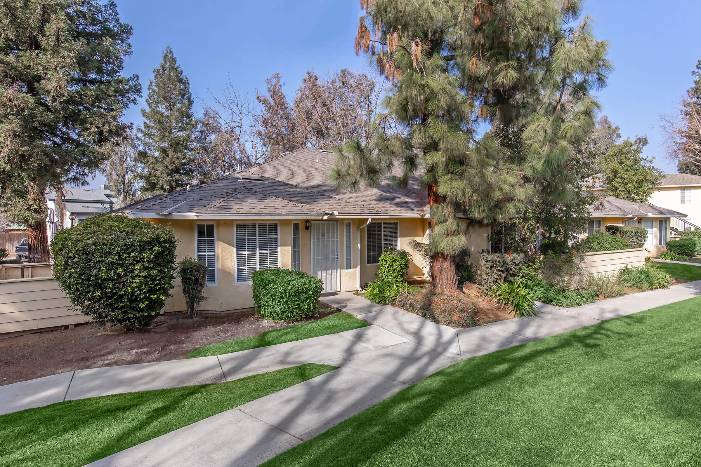 A quaint single-story house surrounded by well-manicured greenery and trees. The home features a welcoming porch with a walkway leading to the entrance. Lush lawns and shrubs frame the property, creating a warm and inviting atmosphere. Clear blue skies complement the charming setting.