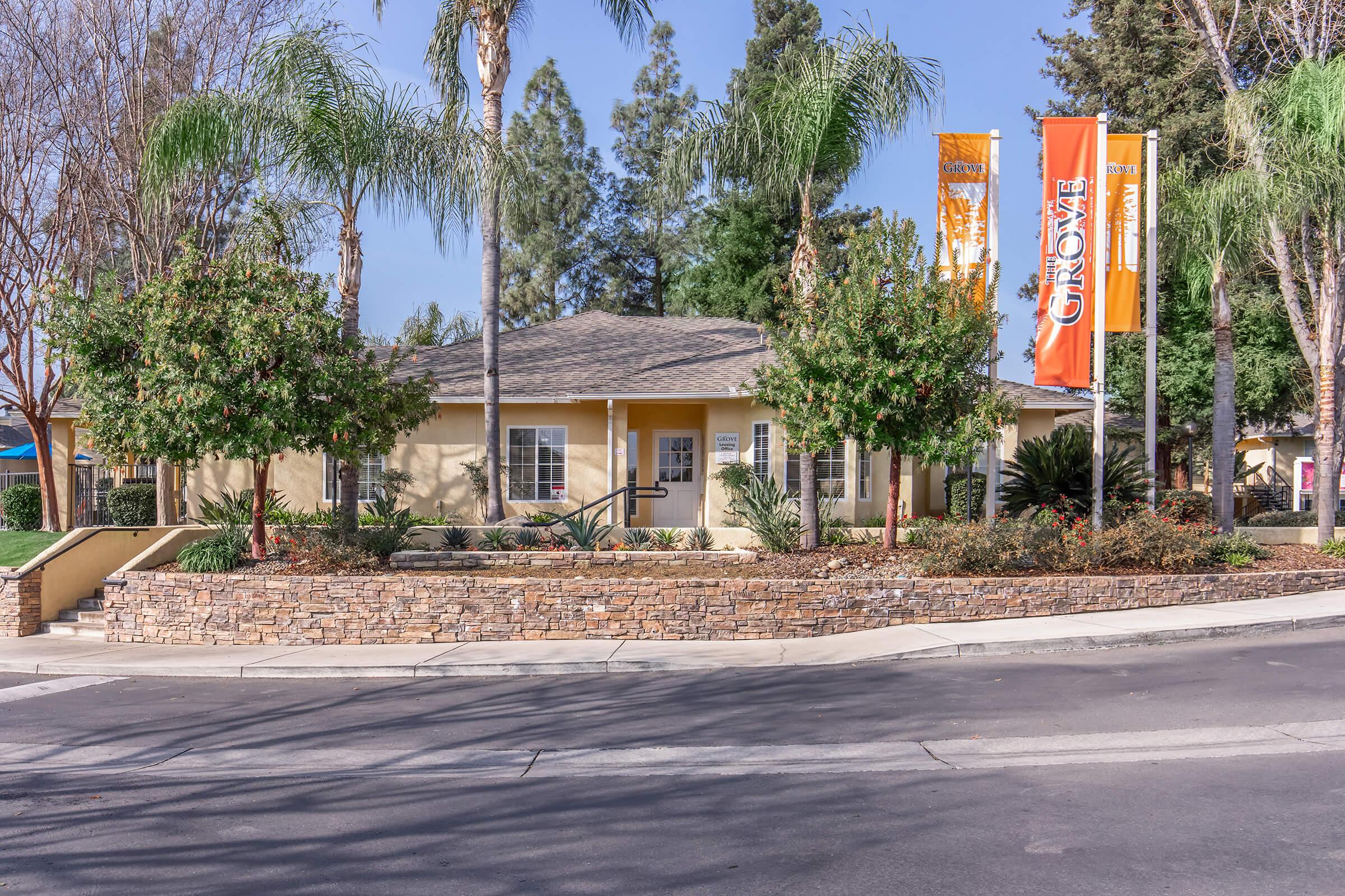 A welcoming building with a sandy-colored exterior, surrounded by lush greenery and palm trees. Two orange banners are displayed in front, and a decorative stone wall outlines the entrance. The scene captures a sunny day, enhancing the inviting atmosphere of the location.