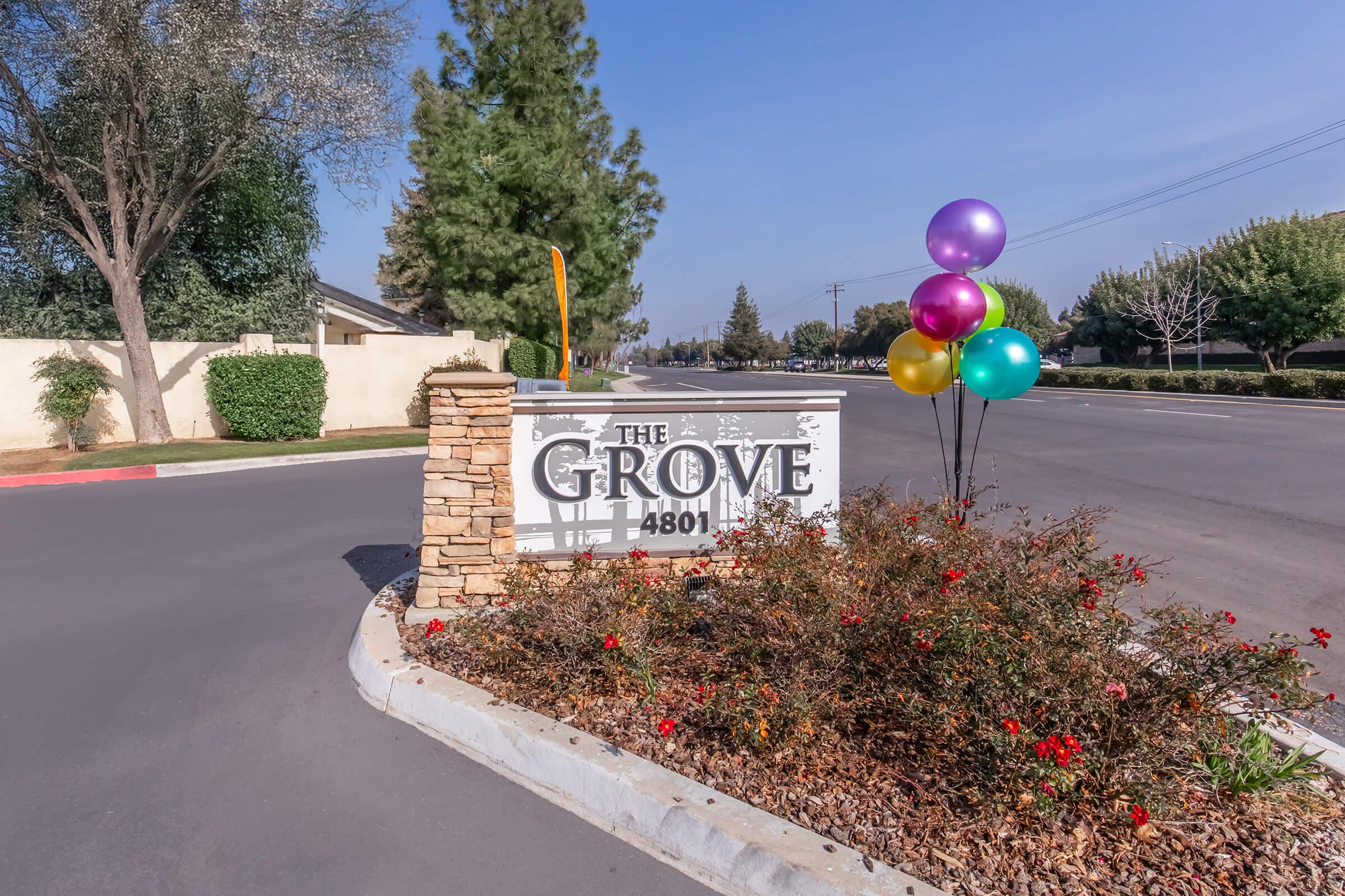 A sign for "The Grove" at 4801, surrounded by colorful balloons and blooming flowers. It is positioned at the entrance of a residential area, with clear skies and trees in the background. The street is visible, suggesting a welcoming atmosphere.