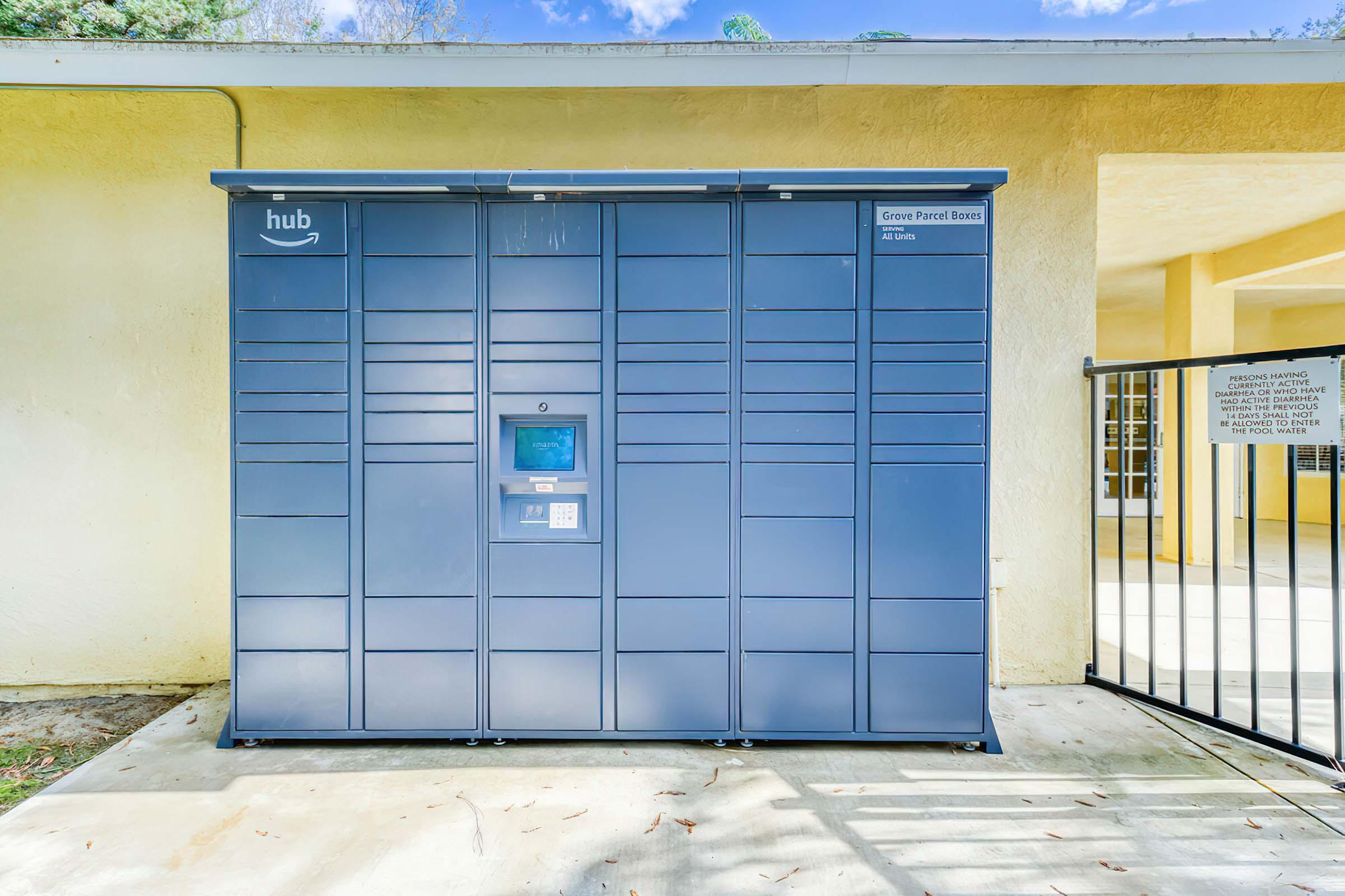 Amazon Hub delivery locker situated outside a building. The locker is blue with the "hub" logo visible. There is a keypad and display screen on the front for user interaction. The surrounding area includes a concrete path and a railing with an entrance to another part of the building in the background.