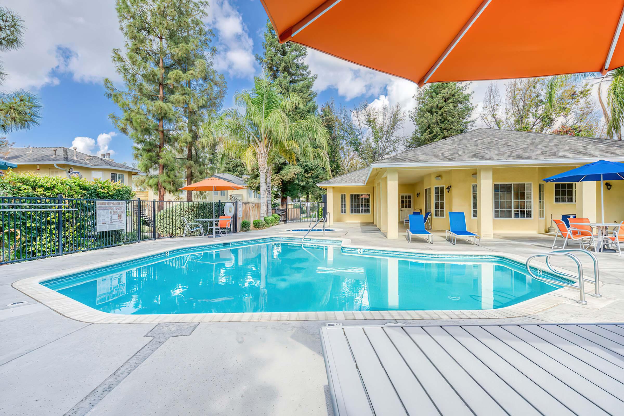 A clear blue swimming pool surrounded by palm trees and lounge chairs, with a bright orange umbrella providing shade. The pool area features a fenced perimeter and a nearby yellow building with multiple windows. The sky is partly cloudy, creating a relaxing outdoor atmosphere.