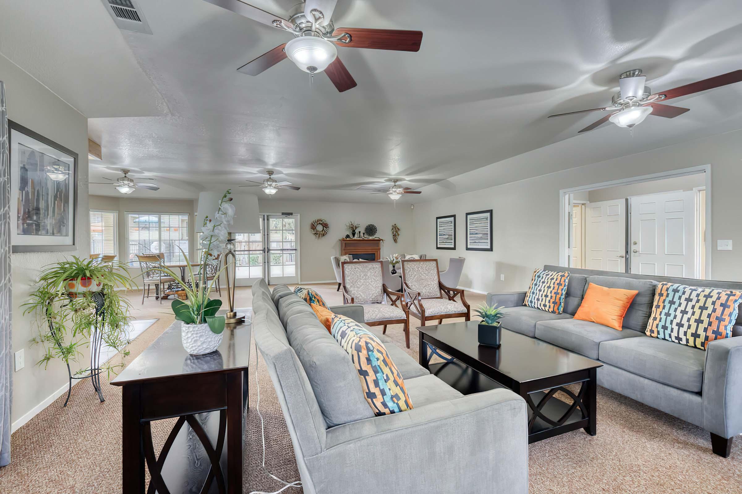Spacious and well-lit living room featuring a comfortable gray sofa with colorful pillows, two wooden armchairs, a coffee table, and potted plants. Ceiling fans are visible above, with natural light coming from large windows. The adjacent dining area is partially visible in the background.