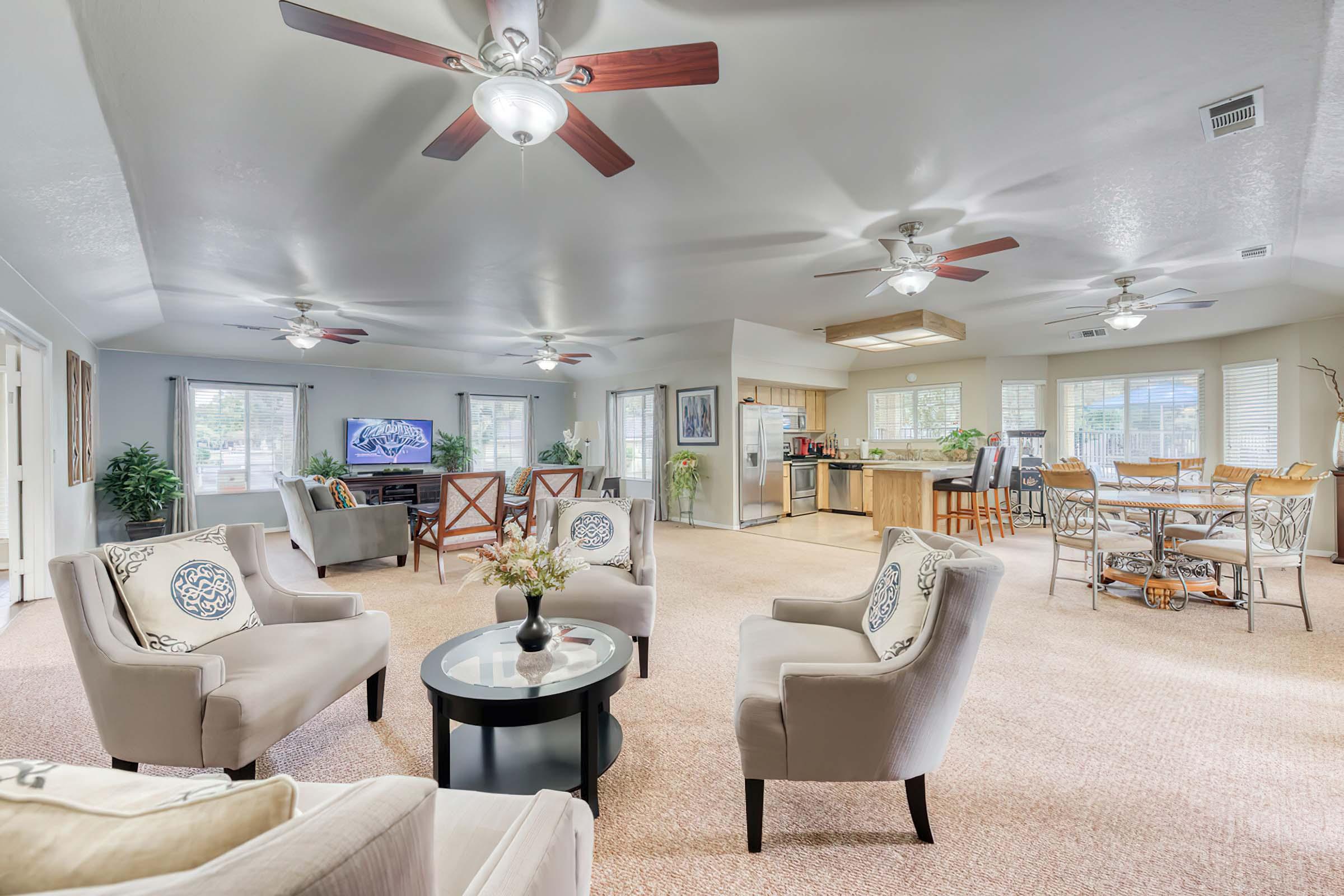 Spacious living room with comfortable seating, light-colored furniture, and decorative pillows. The area features ceiling fans, a coffee table with flowers, and large windows for natural light. An open kitchen is visible in the background, along with a dining area furnished with a round table and chairs.