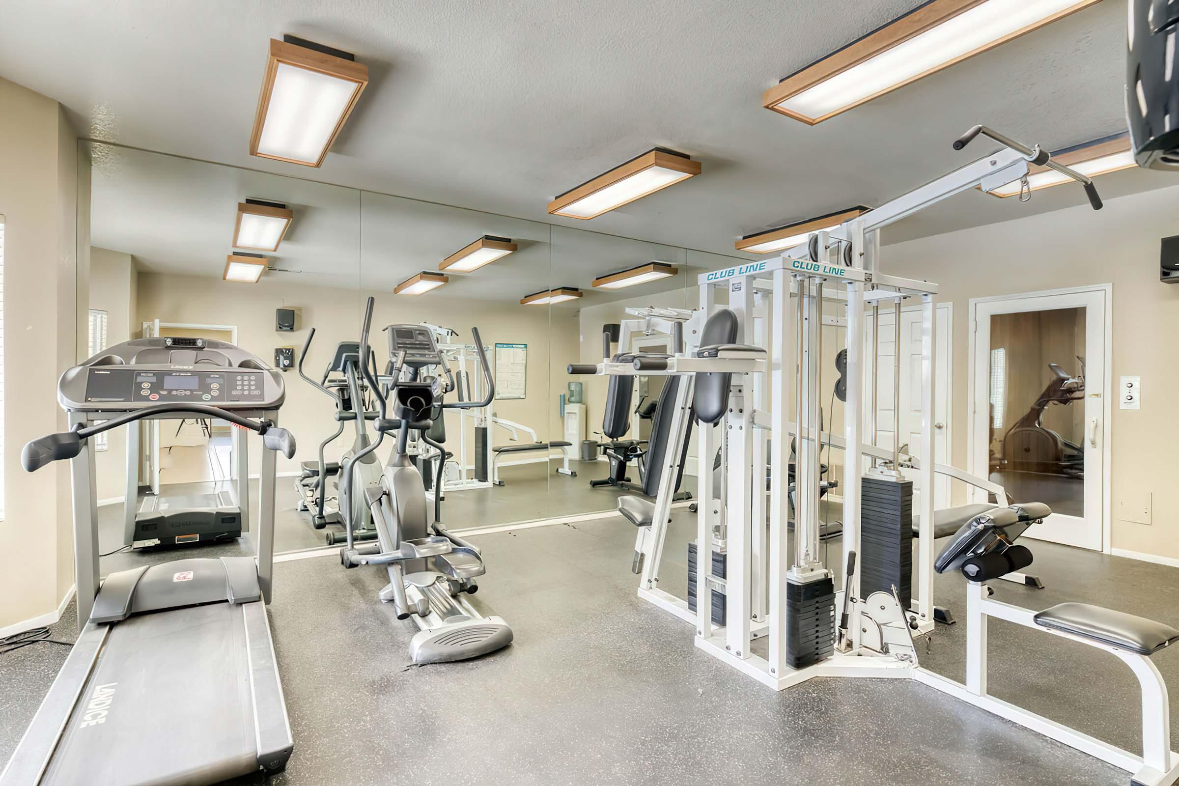 A well-lit gym featuring a treadmill, elliptical machine, and multi-station weight equipment. The space has mirrors on one wall and overhead lighting, creating a bright and inviting atmosphere for exercise. The flooring is dark, complementing the modern fitness equipment.