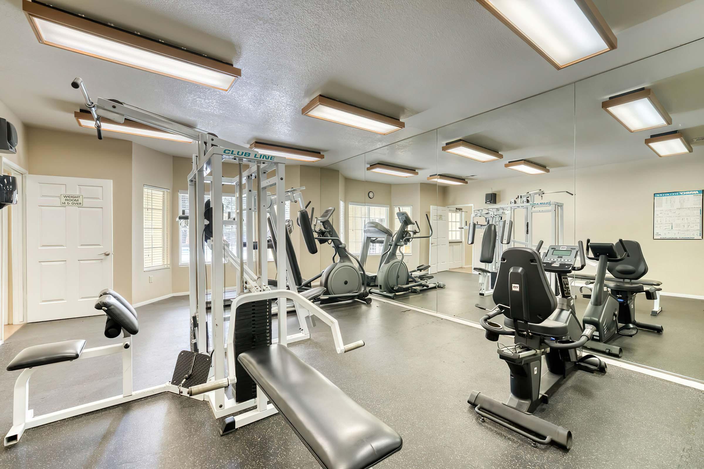 A well-equipped gym featuring exercise machines, weight benches, and cardio equipment. The space is brightly lit with ceiling lights, mirrors on the walls, and large windows allowing natural light. The floor is dark and has a professional finish, creating a welcoming fitness environment.
