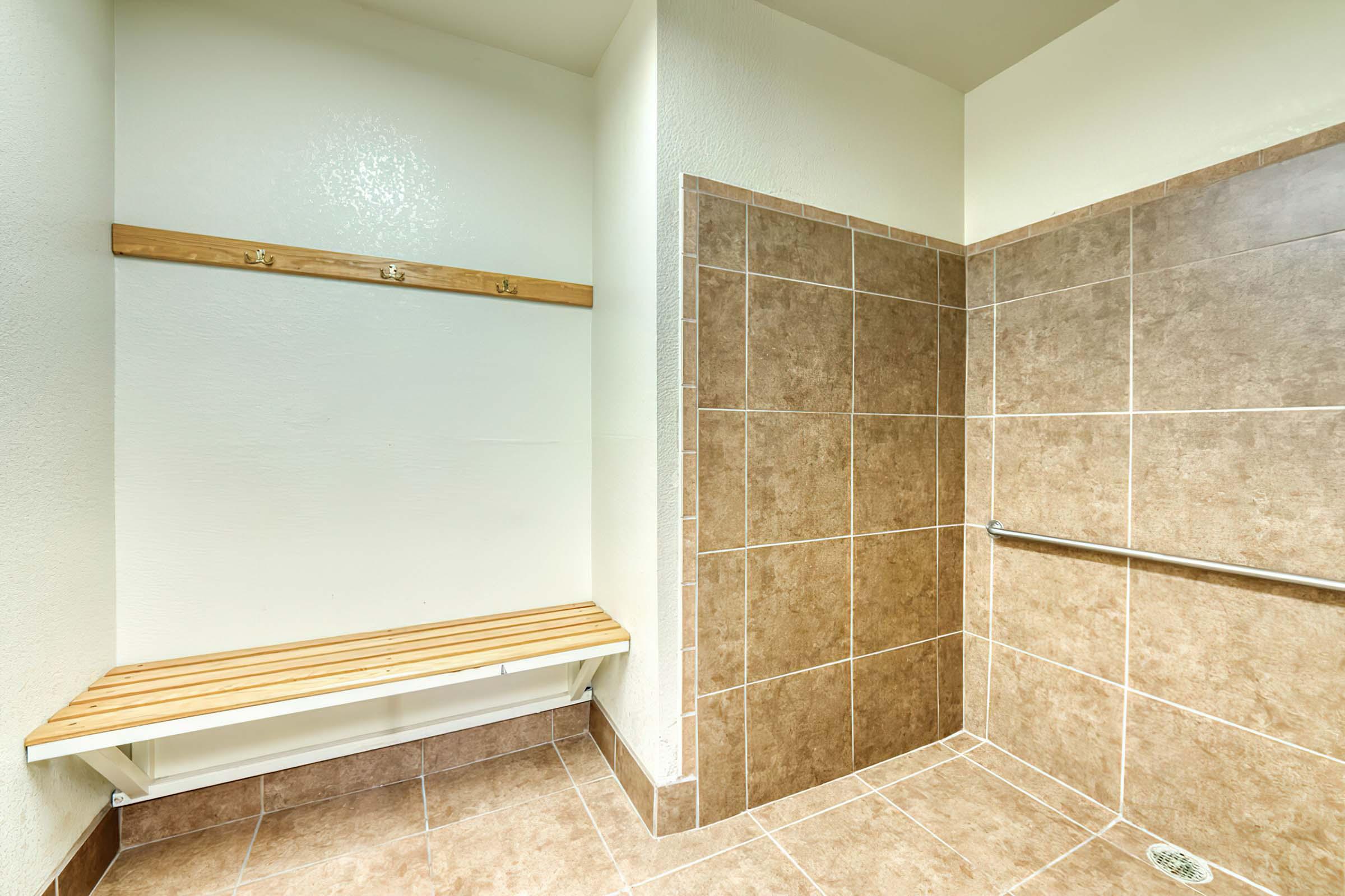 Interior of a clean, modern shower room featuring beige tiled walls and floor. A wooden bench is mounted against one wall beneath coat hooks, and a silver grab bar is installed near the shower area. The space is well-lit and designed for accessibility and comfort.