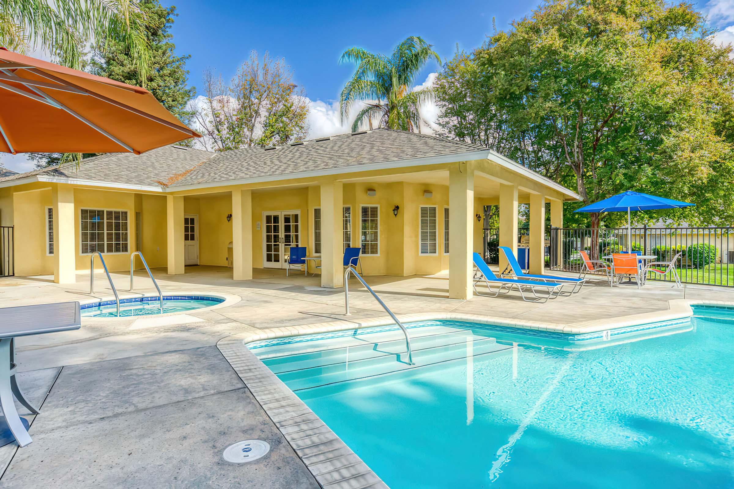 A bright outdoor pool area featuring a swimming pool and a hot tub, surrounded by lounge chairs and umbrellas. The house nearby has large windows and a patio, set against a backdrop of green trees and a clear blue sky.