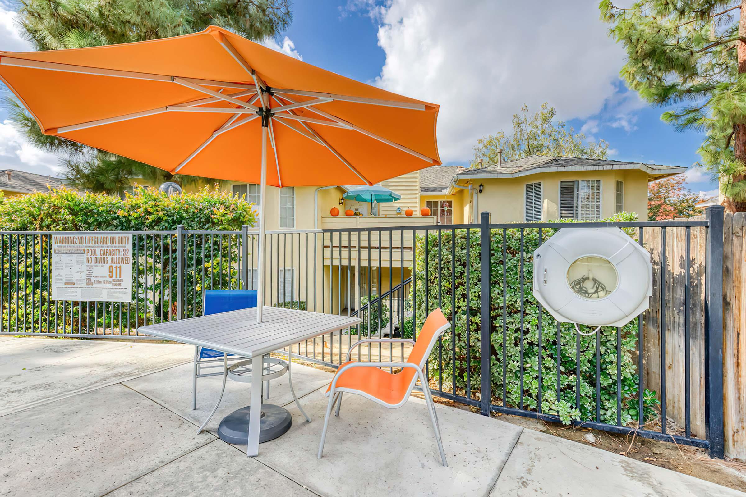 A sunlit outdoor area featuring a table with an orange umbrella and orange chairs, surrounded by greenery. In the background, there is a building with a balcony. A life ring is mounted on a fence, and a poster is visible nearby. The sky is partly cloudy.