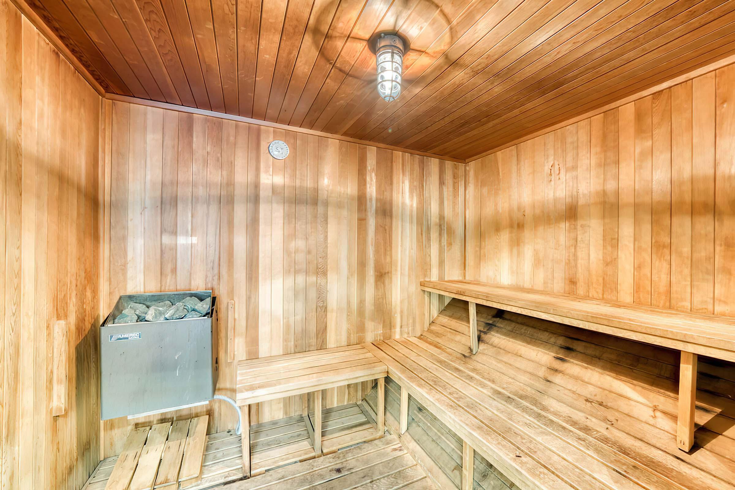 A wooden sauna interior featuring vertical wooden paneling, a stone heater with a metal casing, two wooden benches, and a round clock on the wall. Soft lighting illuminates the space, creating a warm and inviting atmosphere.