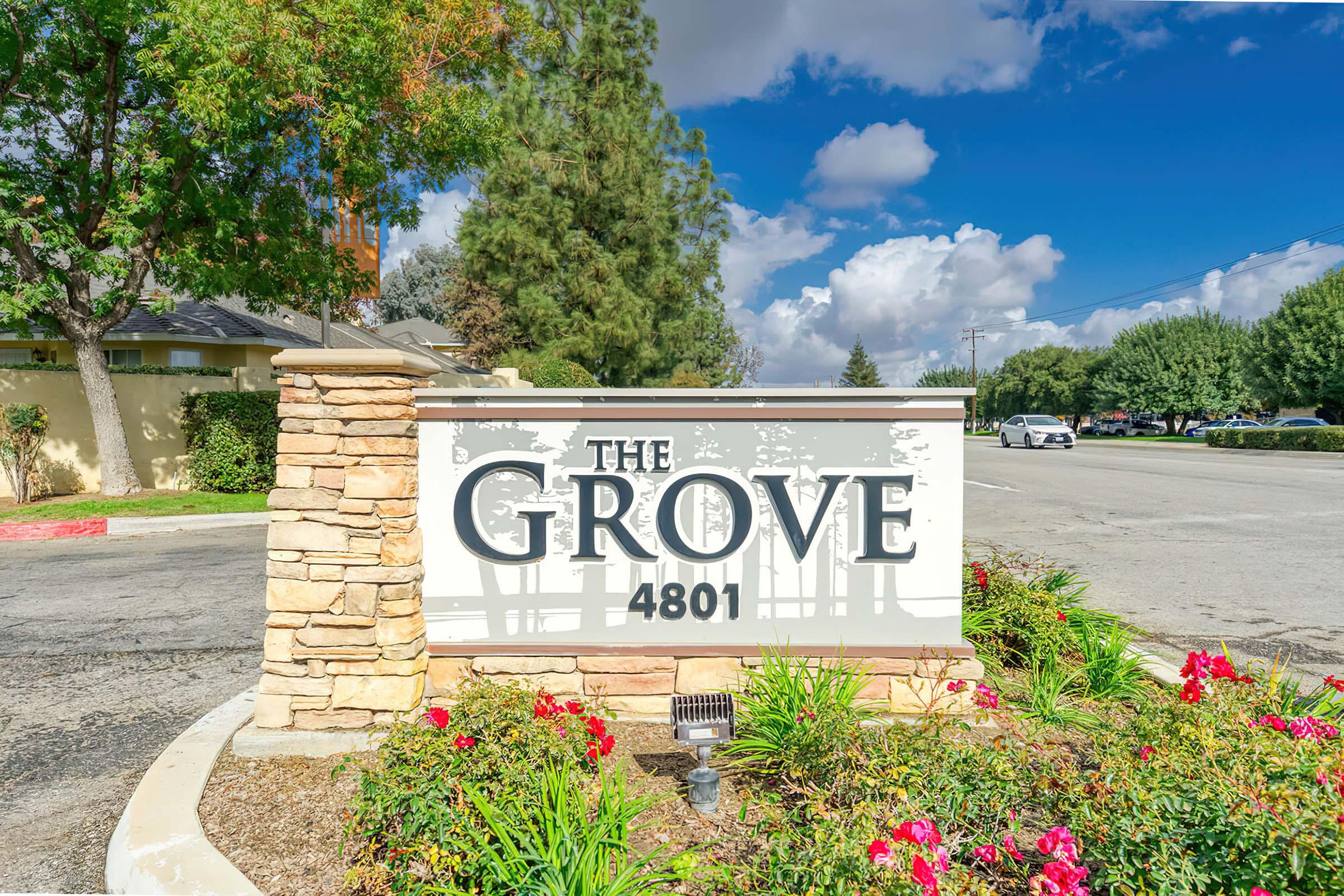 Sign for "The Grove" located at 4801, featuring a stone base and surrounded by colorful flowers. In the background, blue skies with clouds and green trees can be seen along the road.