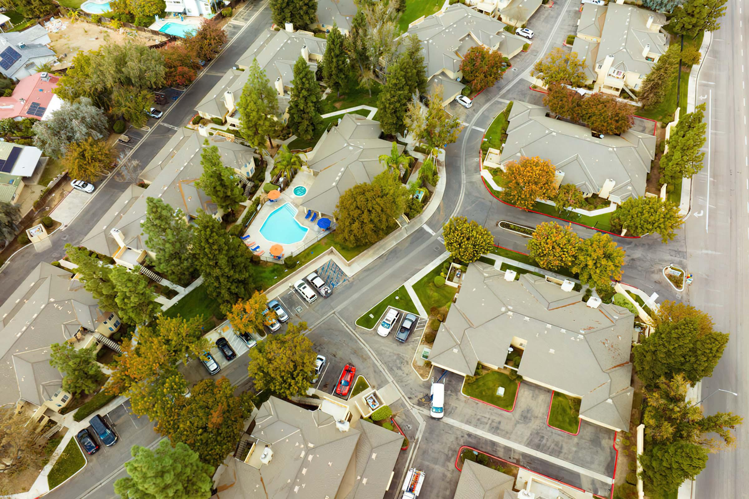 Aerial view of a suburban neighborhood featuring several houses with sloped roofs, a swimming pool in one yard, and colorful trees, with cars parked along the streets. The layout includes winding roads and green spaces, creating a picturesque residential area.