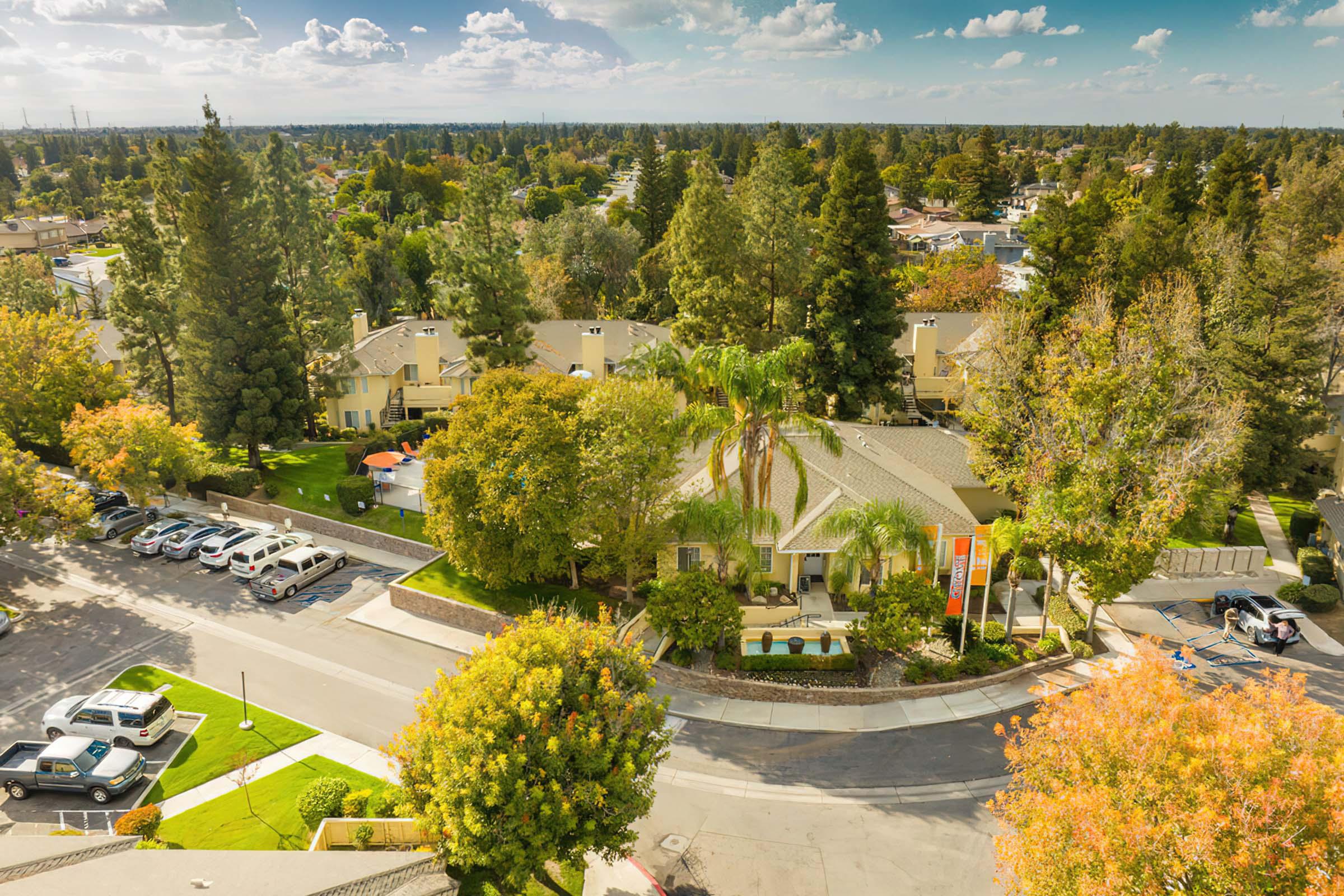Aerial view of a suburban neighborhood featuring green trees, landscaped lawns, and several residential buildings. A circular street is visible, with parked cars along the curb. The scene captures a mix of greenery and residential architecture, indicative of a quiet, well-maintained community.