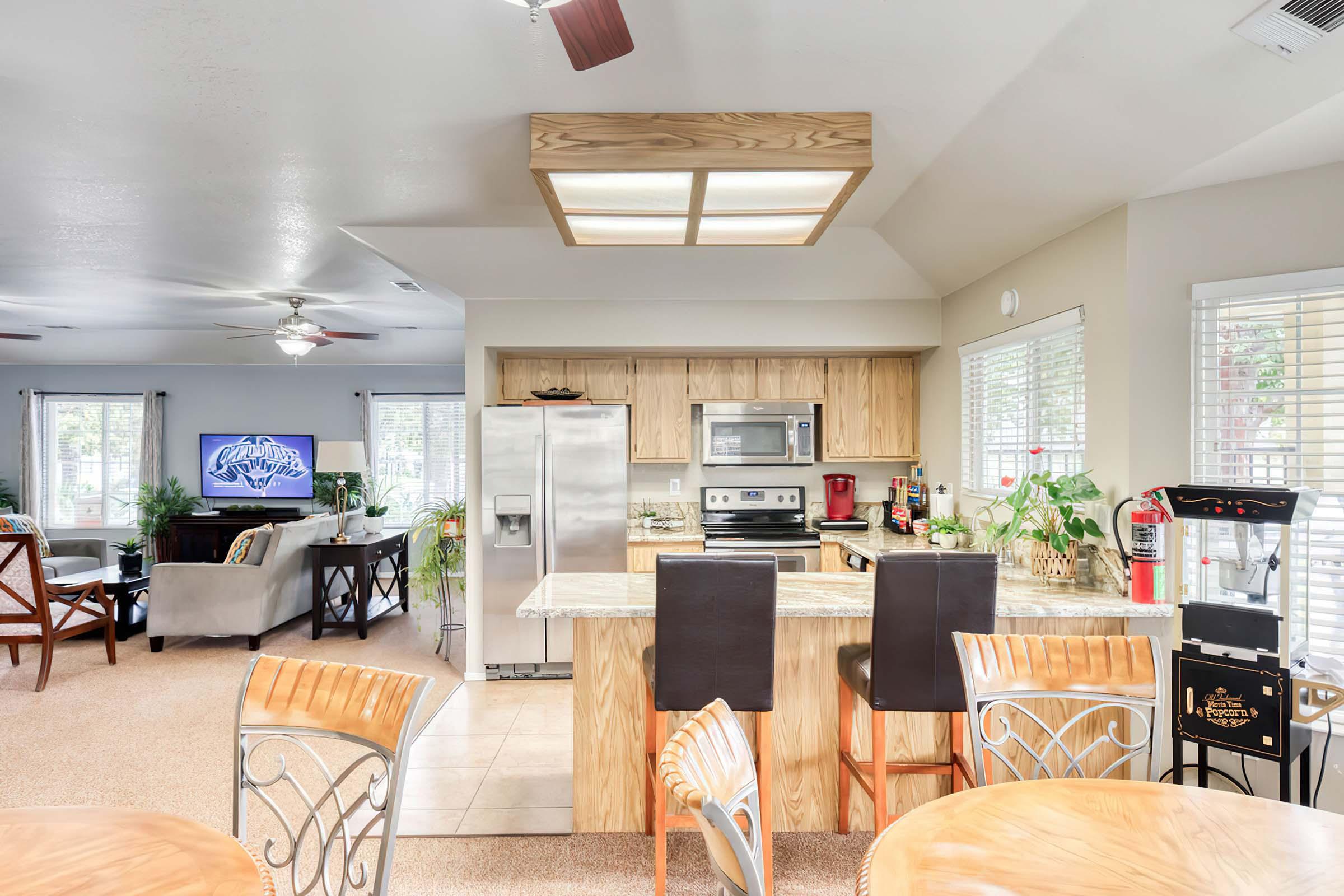 A modern kitchen featuring a central island with bar stools, stainless steel appliances, and wooden cabinets. There is a cozy living area visible in the background with a television and comfortable seating. Natural light flows in through large windows, and indoor plants add a touch of greenery.