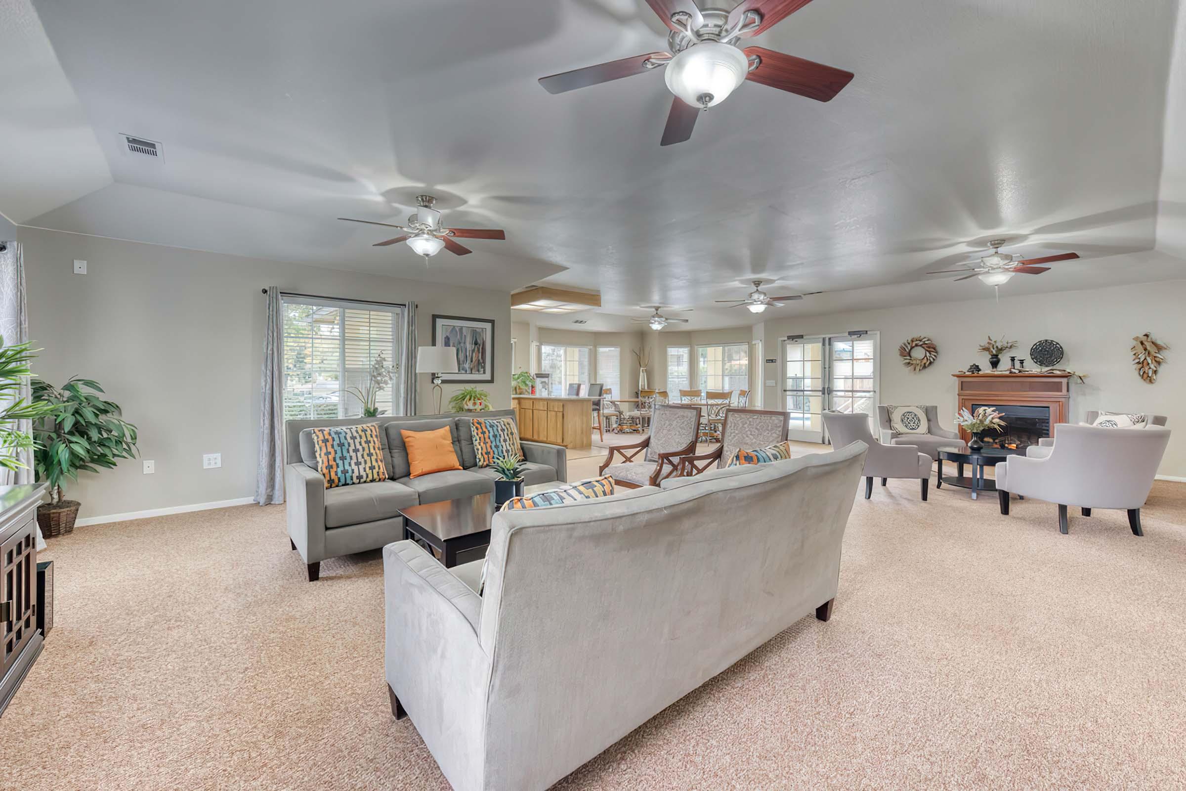 A spacious living room featuring a light-colored sofa with decorative pillows, two comfortable armchairs, and a coffee table. Ceiling fans illuminate the room, which has a carpeted floor and large windows allowing natural light. A dining area is visible in the background, along with decorative elements on the walls.