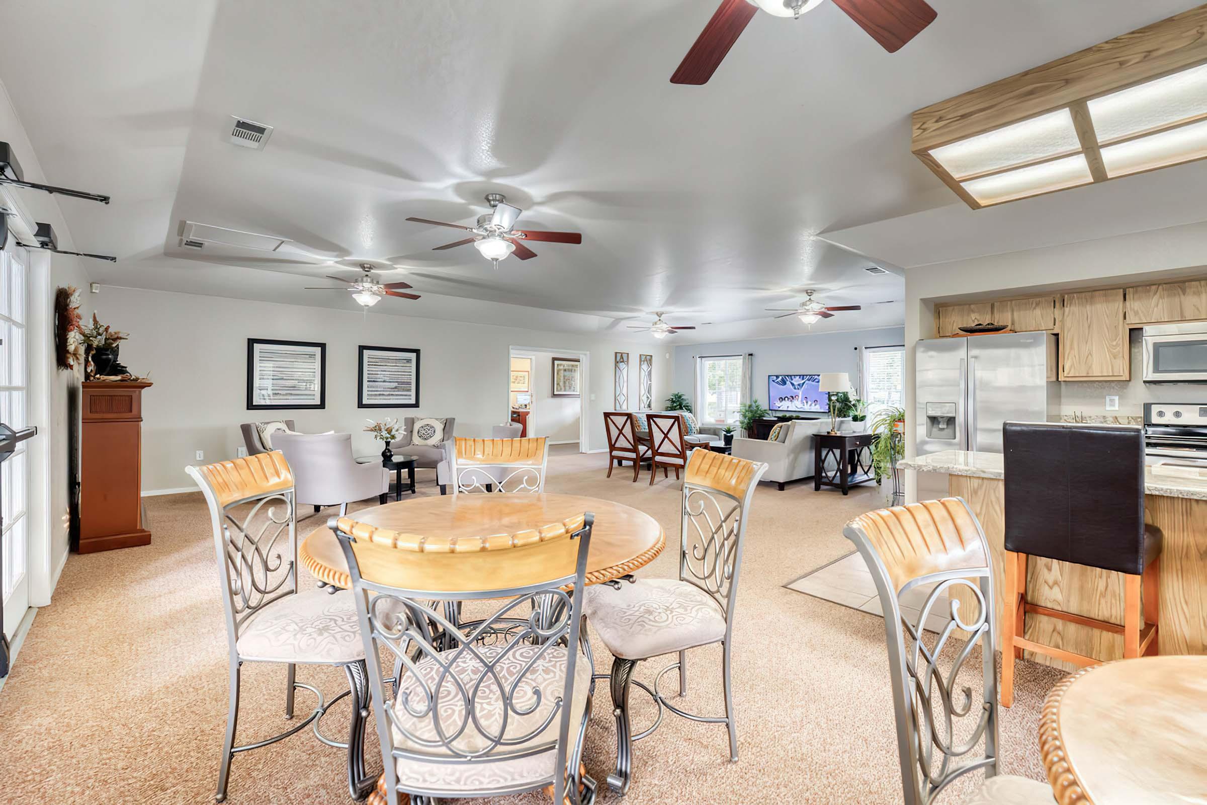 A spacious, well-lit living area featuring a dining table with decorative chairs, a cozy seating arrangement, ceiling fans, and large windows. The open layout includes a kitchen area with modern appliances and a neutral color palette, creating a warm and inviting atmosphere.