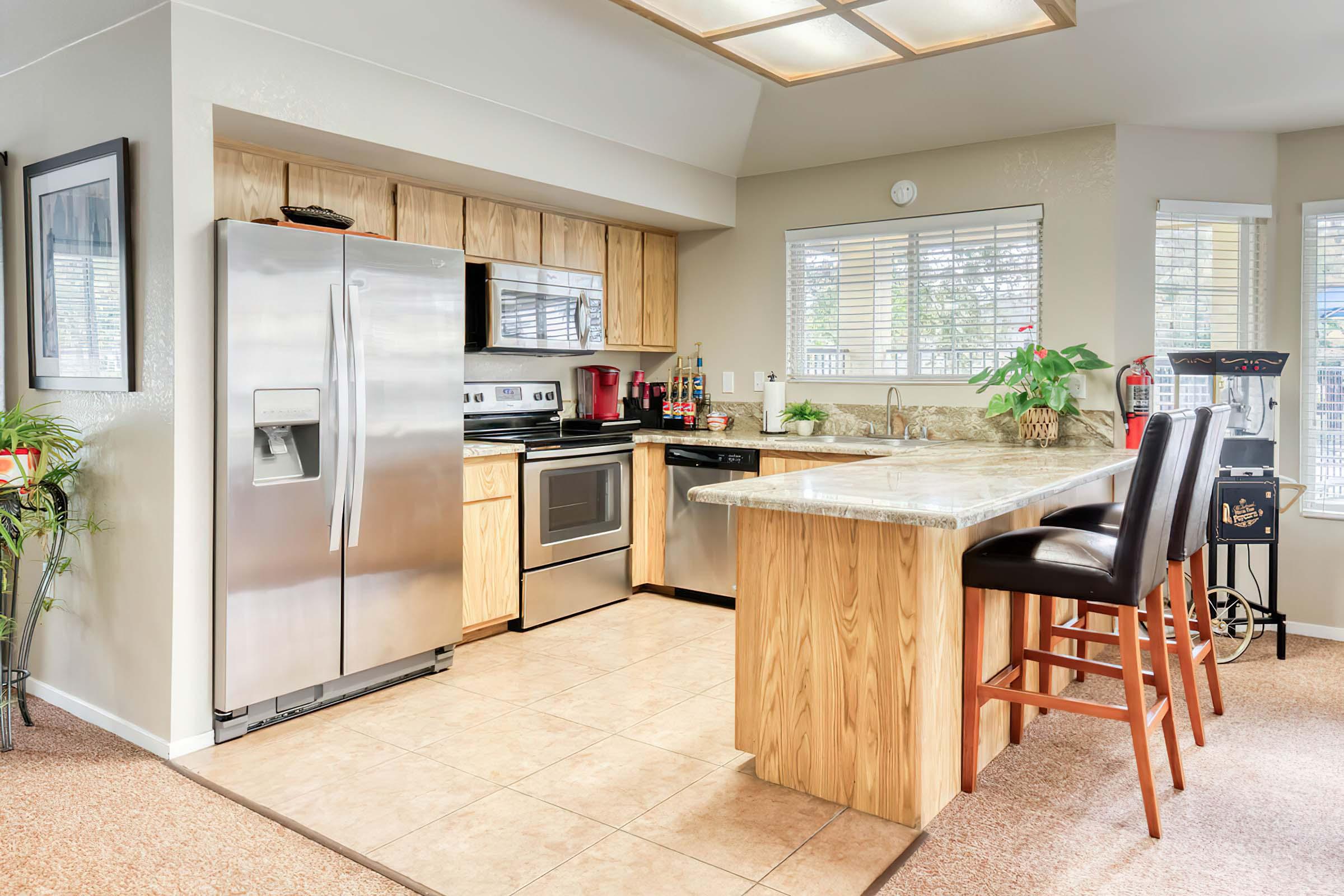 Modern kitchen featuring stainless steel appliances, wood cabinetry, and a marble countertop. The island has three bar stools, and there are windows providing natural light. Decorative plants and kitchenware add a cozy touch.