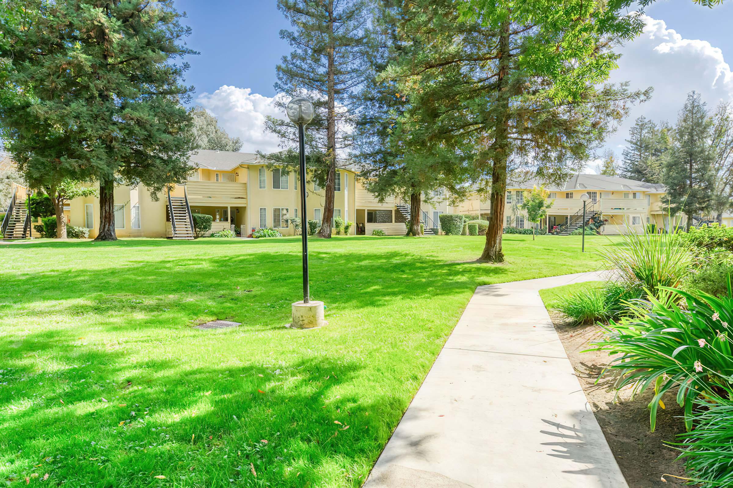 A well-maintained grassy area featuring several tall trees. A concrete path winds through the lawn, leading to a series of apartment buildings with light-colored siding. A lamp post stands along the path, and the sky is bright with fluffy clouds.