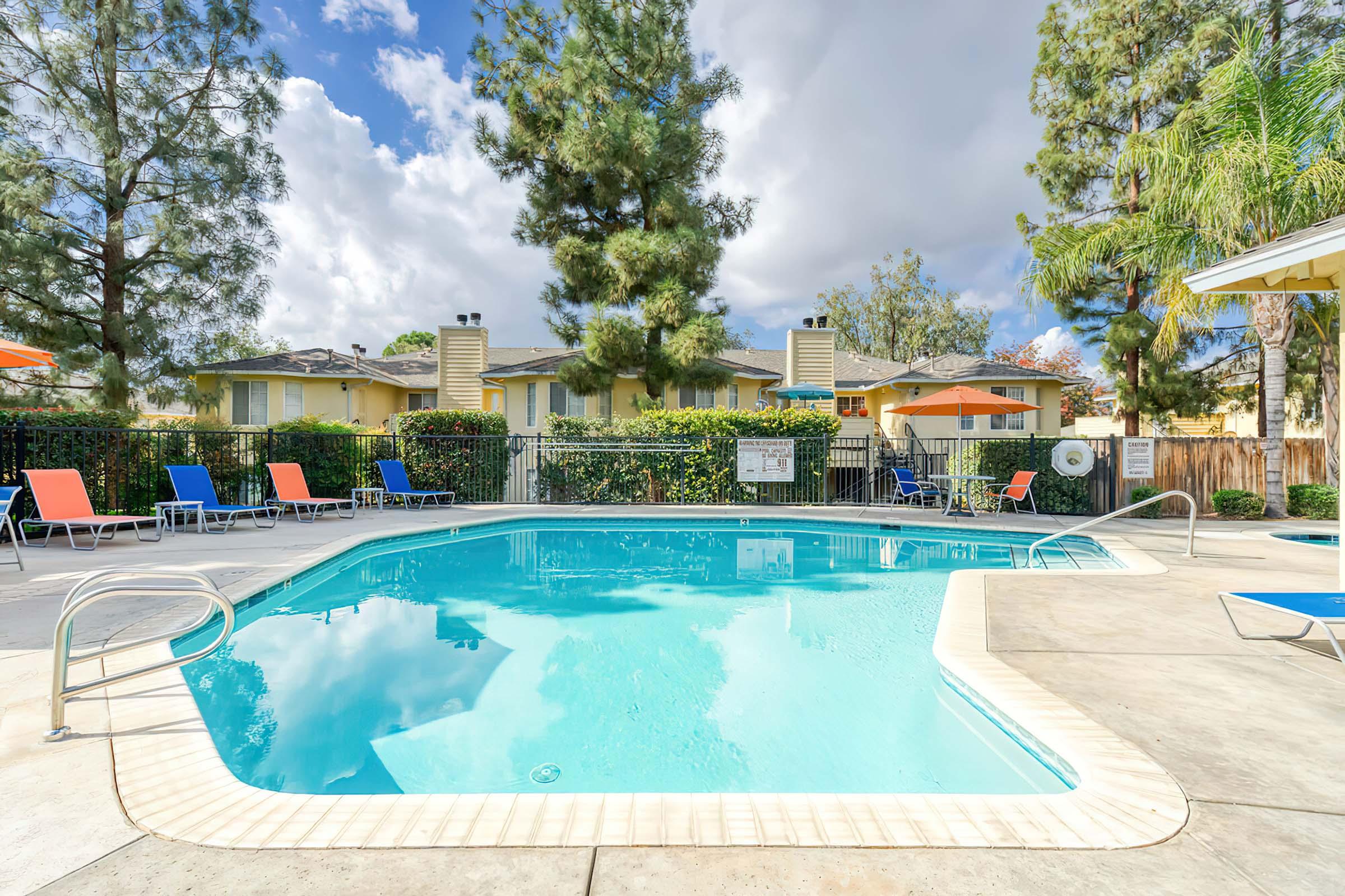A clean, inviting swimming pool surrounded by lounge chairs in a residential area. Bright blue water reflects the sky, with fluffy clouds overhead. Colorful umbrellas provide shade, while well-maintained greenery frames the scene, creating a relaxing outdoor atmosphere.