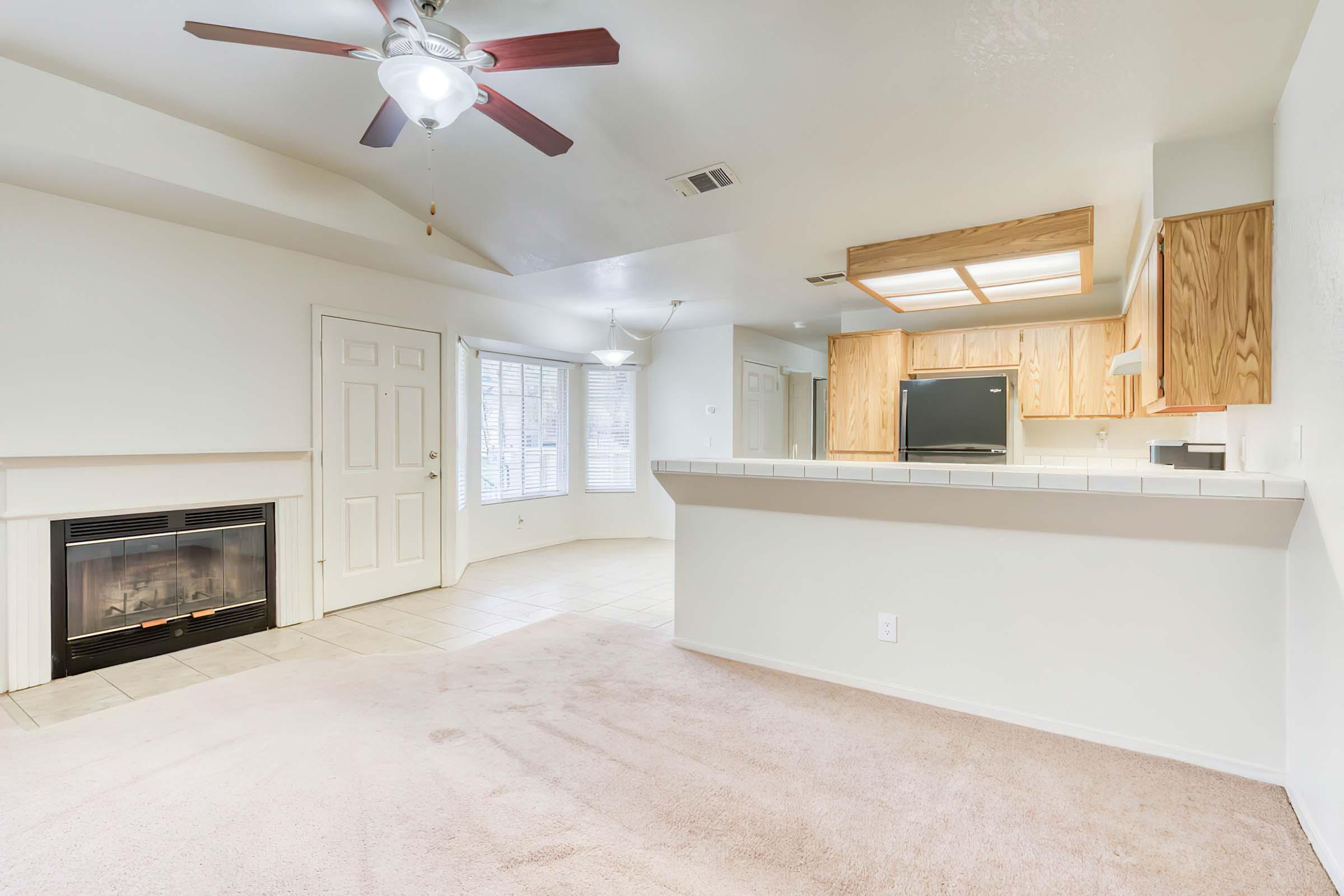 A spacious living area with light-colored walls and carpet, featuring a ceiling fan and a fireplace. There is a small kitchen area with wooden cabinets and a counter that overlooks the living space. Natural light enters through windows, creating a bright and inviting atmosphere.