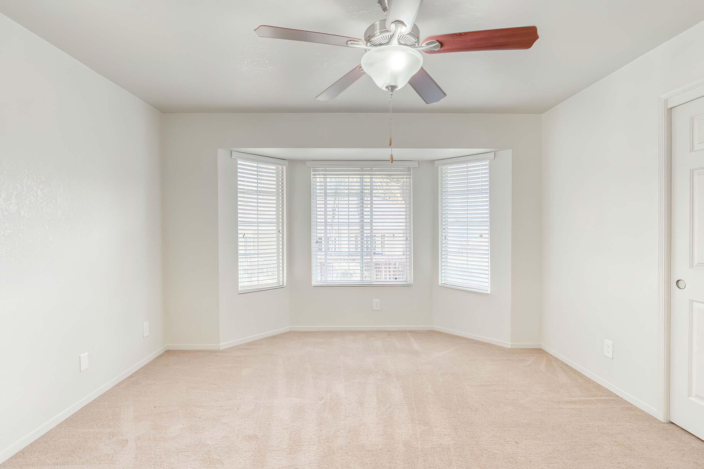 A spacious, empty room with beige carpeting and white walls. It features a ceiling fan with wooden blades, and a large bay window with white blinds that allows natural light to fill the space. A white door is visible on the right side of the image.