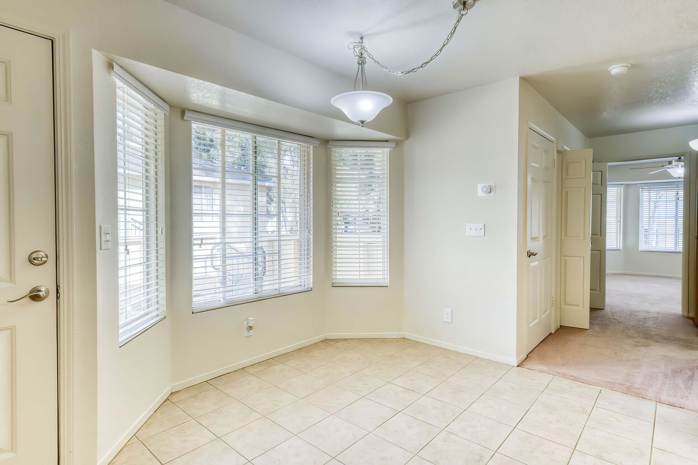 Spacious interior view of a well-lit room featuring a bay window with blinds, beige walls, and tiled flooring. A ceiling light fixture is visible, providing illumination. A doorway leads to another room, enhancing the open layout of the space.