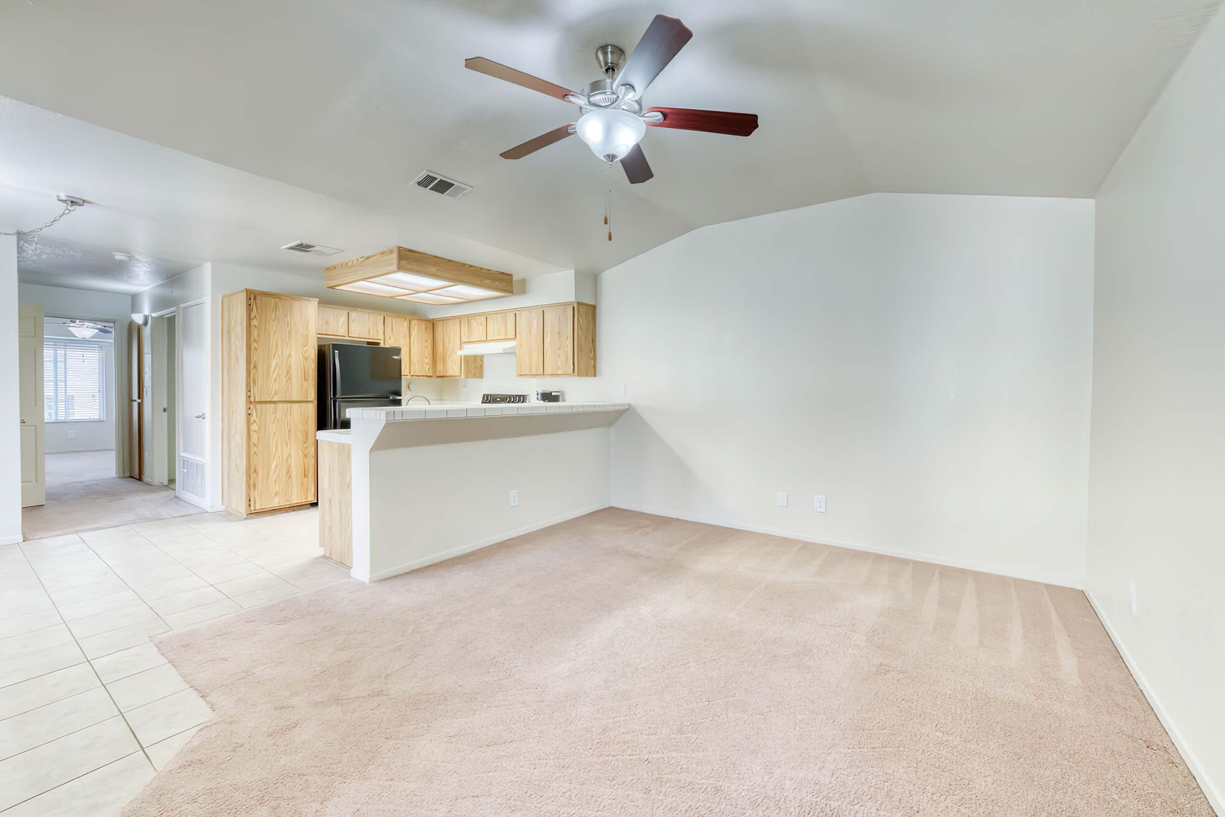 Spacious living area with light-colored walls and beige carpet. A ceiling fan is present above, and the kitchen area features wooden cabinets and a countertop. Open layout leading to a hallway, with doorways visible on the left. Natural light brightens the room through a window.
