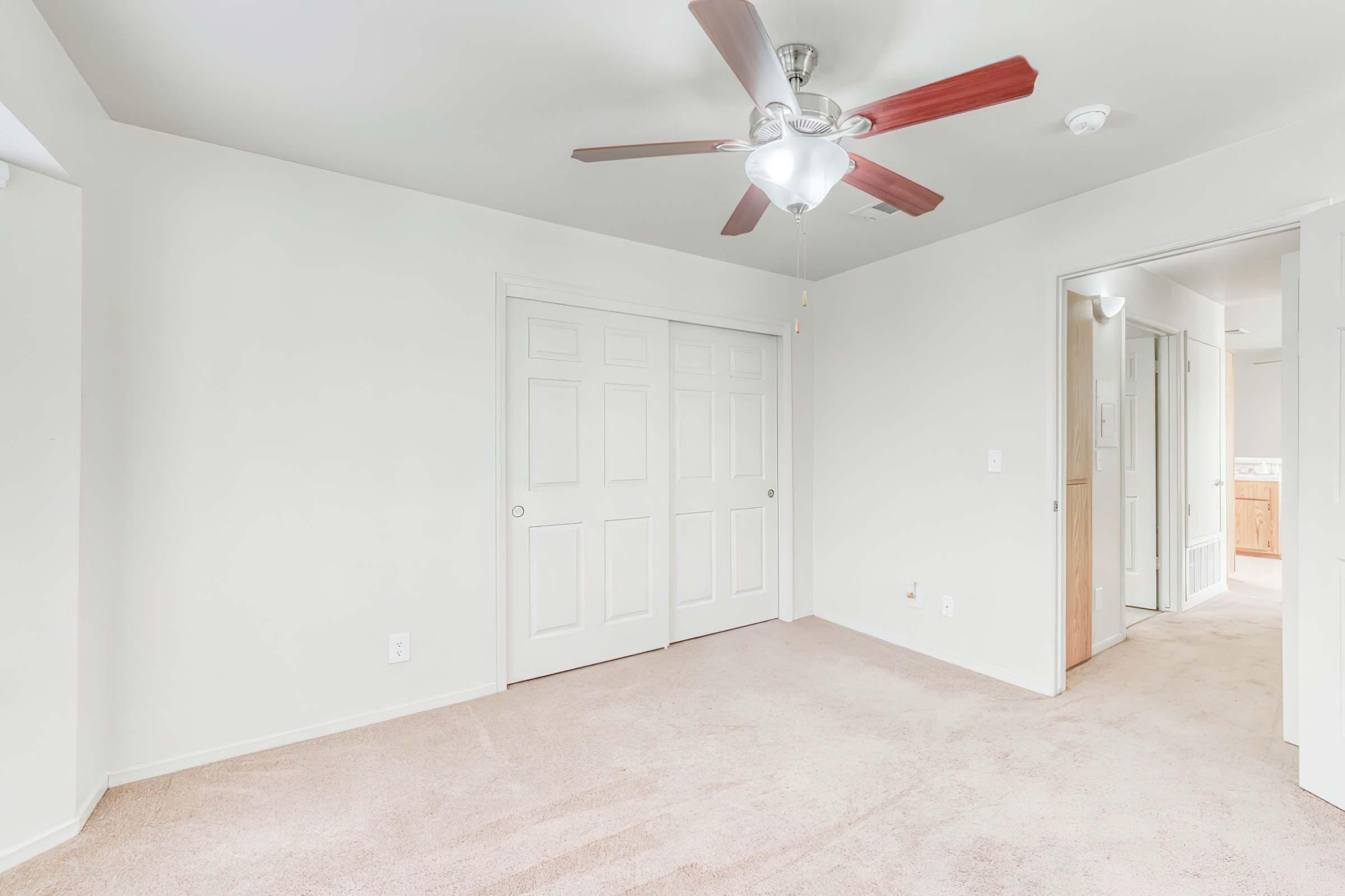 A well-lit, empty bedroom featuring light beige carpet and white walls. It includes a ceiling fan with wooden blades, double doors for a closet, and an adjacent doorway leading to a bathroom area. The space appears airy and inviting, suitable for personal customization.