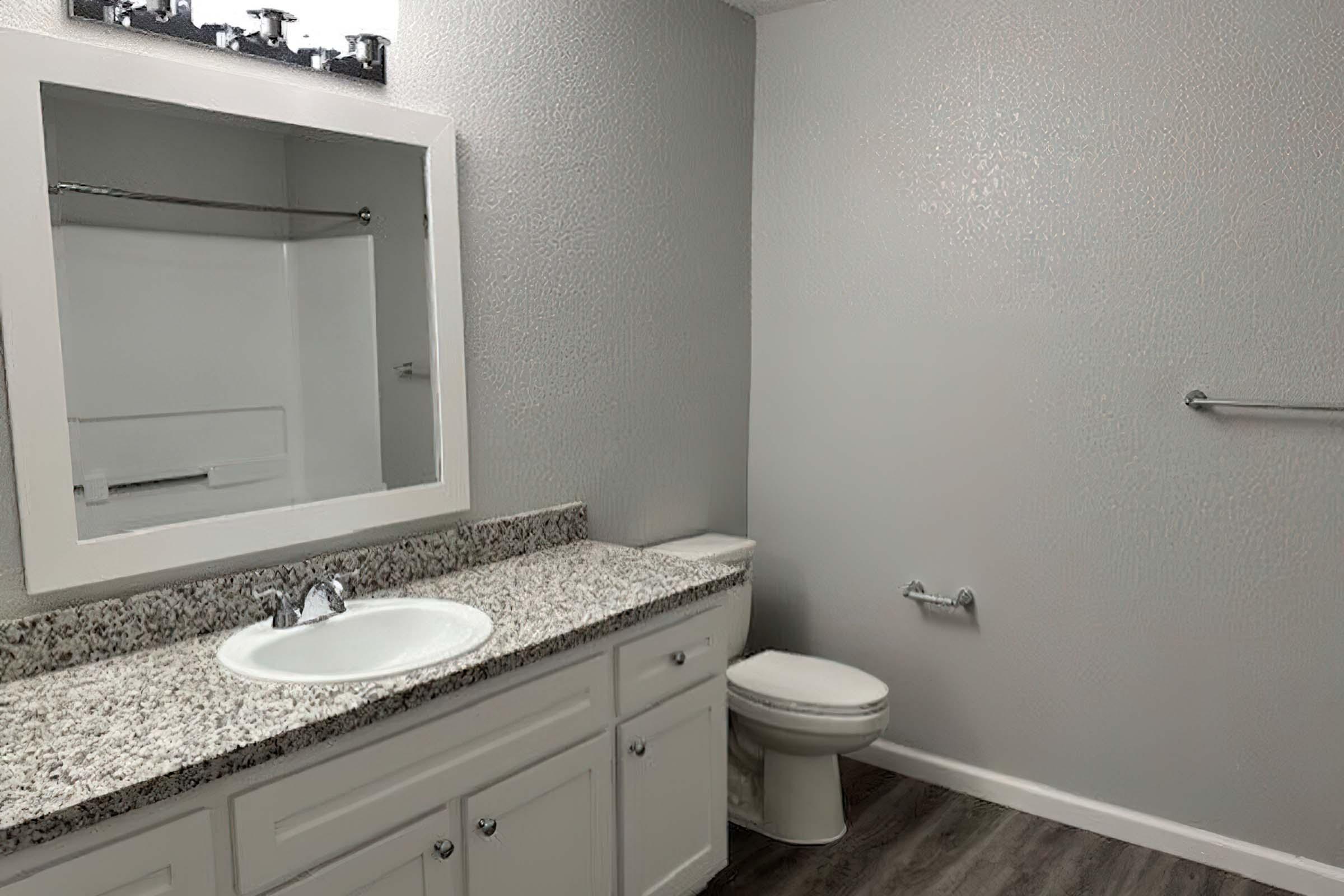 A modern, clean bathroom featuring a white countertop with a sink, a large mirror above it, a toilet, and simple gray walls. The floor is made of wood-like tiles. There are no decorative elements, giving it a minimalist look.