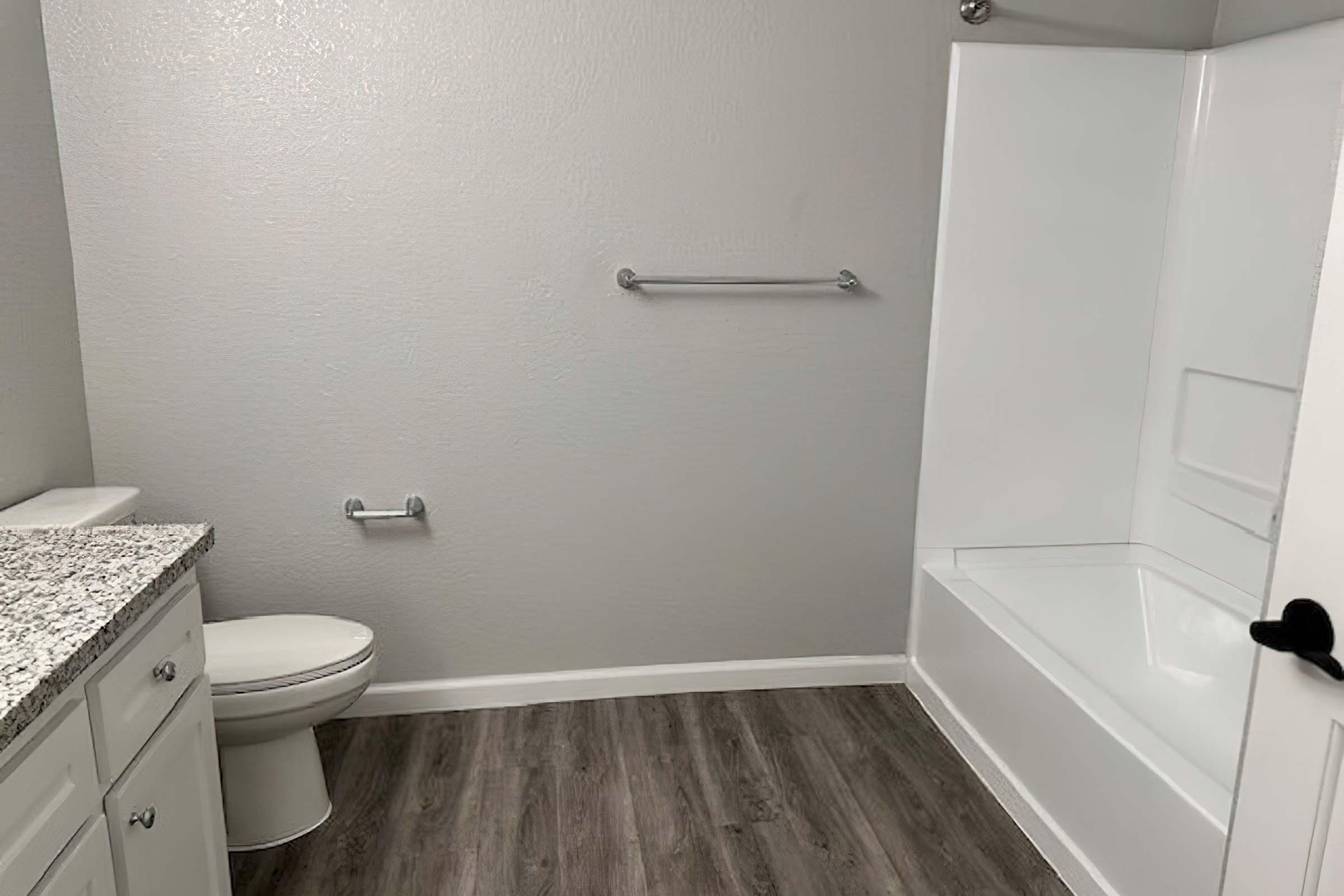 A clean, modern bathroom featuring a white bathtub, a toilet, and a granite countertop with cabinets. The walls are painted light gray, and there are simple towel bars mounted on the wall. The flooring is a wood-like laminate, adding to the contemporary look.