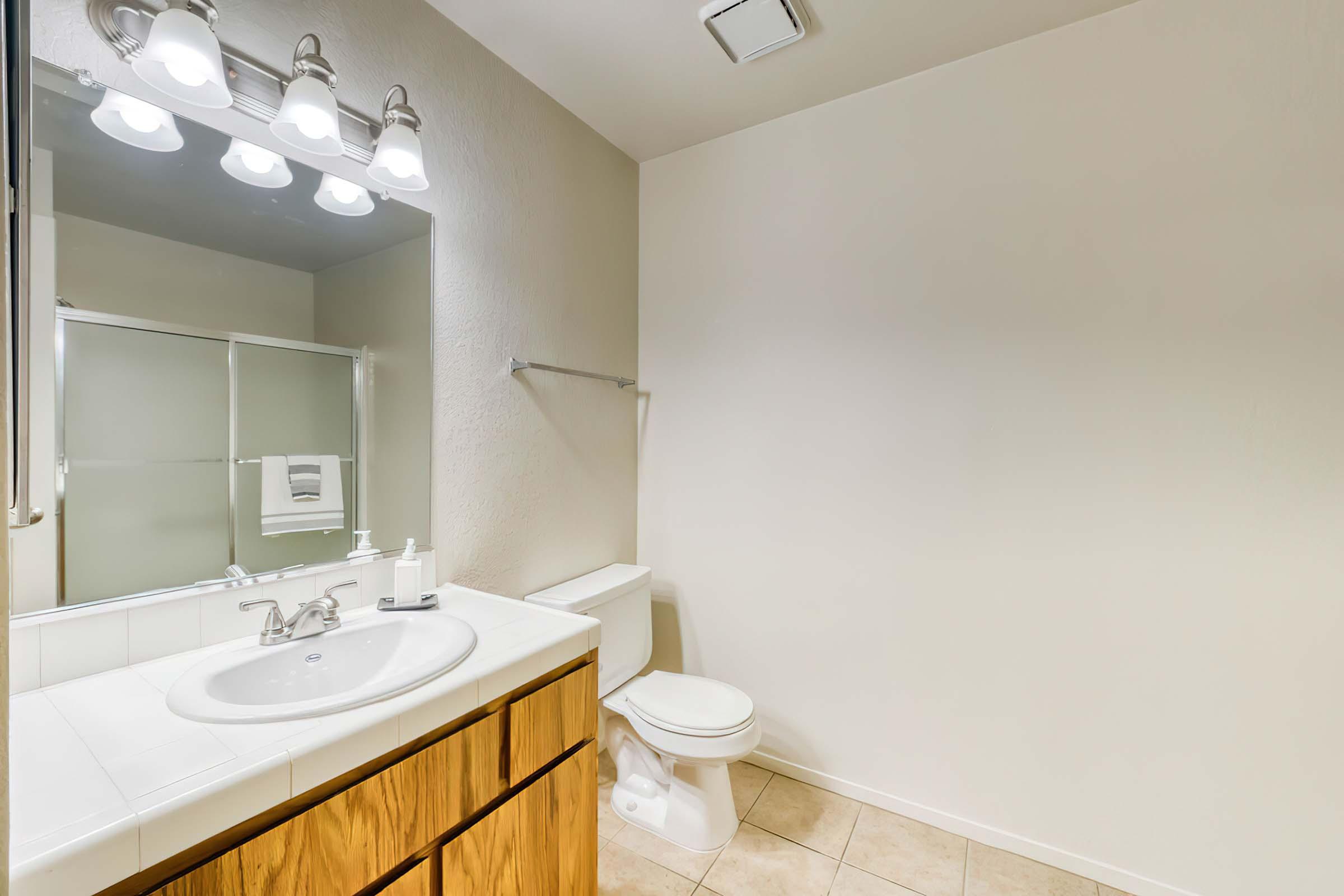 A clean and well-lit bathroom featuring a white sink with a silver faucet, wooden cabinetry, a mirror with four light fixtures above, a toilet nearby, and a glass shower enclosure. The walls are painted a light color, and the floor is tiled in a neutral shade.